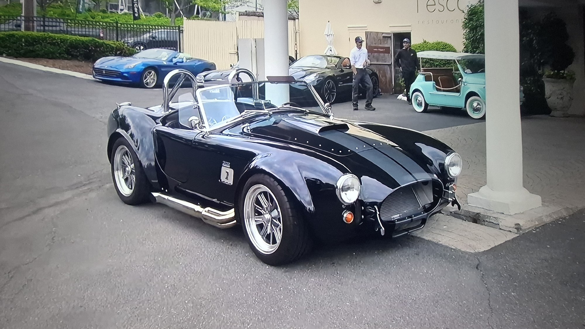 An outdoor, street-level photograph showcases a lineup of distinctive vehicles parked near a building entrance, likely a restaurant. Dominating the foreground is a black AC Cobra-style convertible, featuring two light gray racing stripes, prominent chrome side exhaust pipes, and multi-spoke chrome wheels. A number "7" is visible on its side, and it has a light blue interior.
Behind the Cobra, slightly to the left, a bright blue convertible sports car, possibly a Ferrari, is parked further down the paved area. To the right, beneath a building overhang, another dark-colored luxury convertible with its top down is visible. Adjacent to the building's wooden doors, a charming, vintage-style light blue golf cart with white-walled tires and a white canopy is parked.
Two men are standing near the building's entrance; one wearing a white shirt and baseball cap, the other in dark clothing and a cap. The building itself is light-colored with prominent pillars and wooden doors. A sign above the entrance partially reads "Tesc..." with the word "restaurant" clearly visible below it, indicating the establishment's purpose. Lush green foliage and a black metal fence are in the background.
The scene takes place on a bright day, suggesting pleasant weather suitable for convertibles. The arrangement of high-end and classic cars implies an upscale setting, possibly a car show, valet service, or a gathering of enthusiasts.