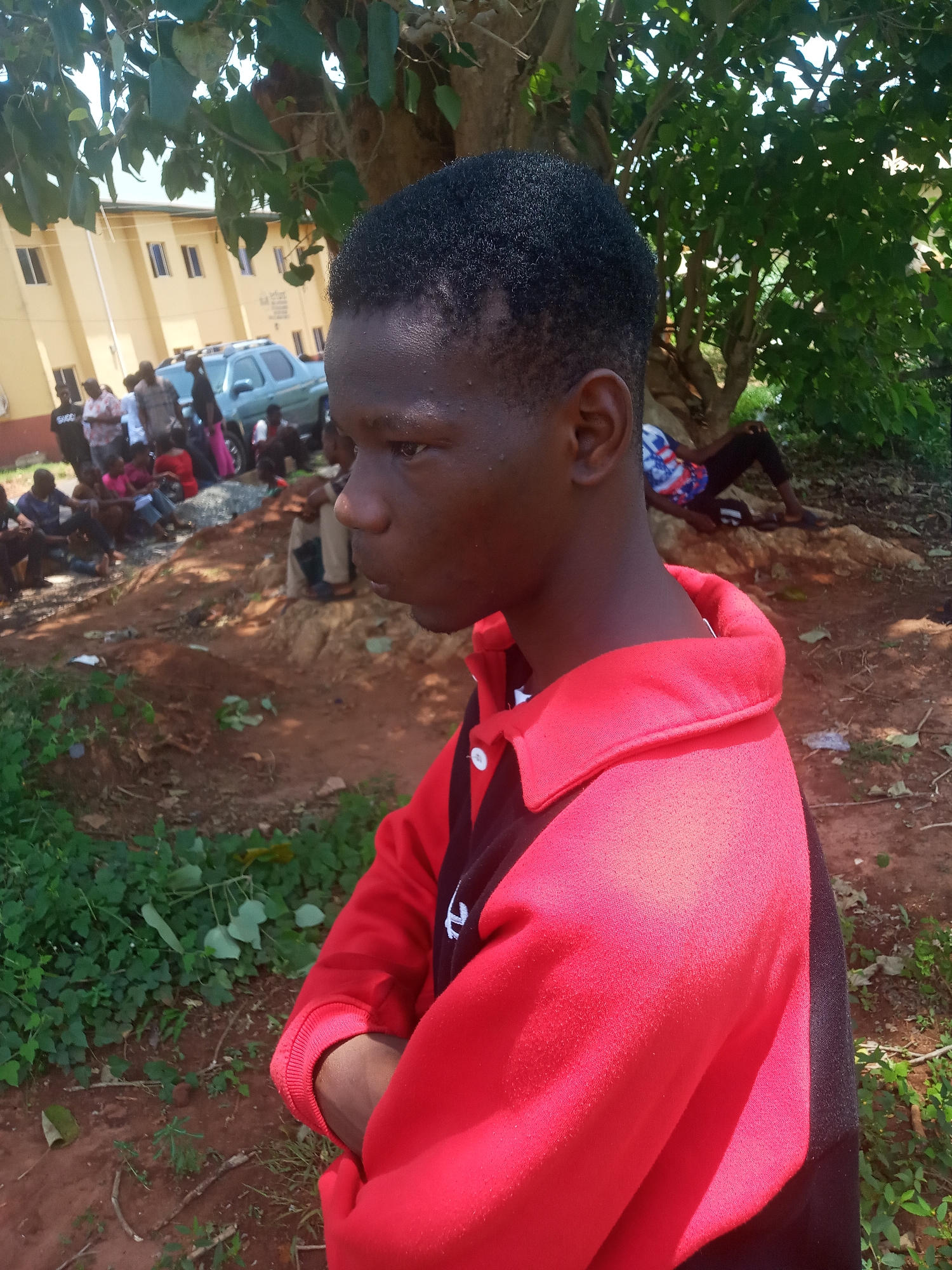 An image shows a young man in a red and black athletic shirt standing with his arms crossed in what appears to be an outdoor area. A group of people are gathered near a building in the background, under the shade of trees. A vehicle can also be seen in the background. The ground around the man is covered with foliage and some bare earth. The location is possibly Ukwu-Abwa, Nigeria.