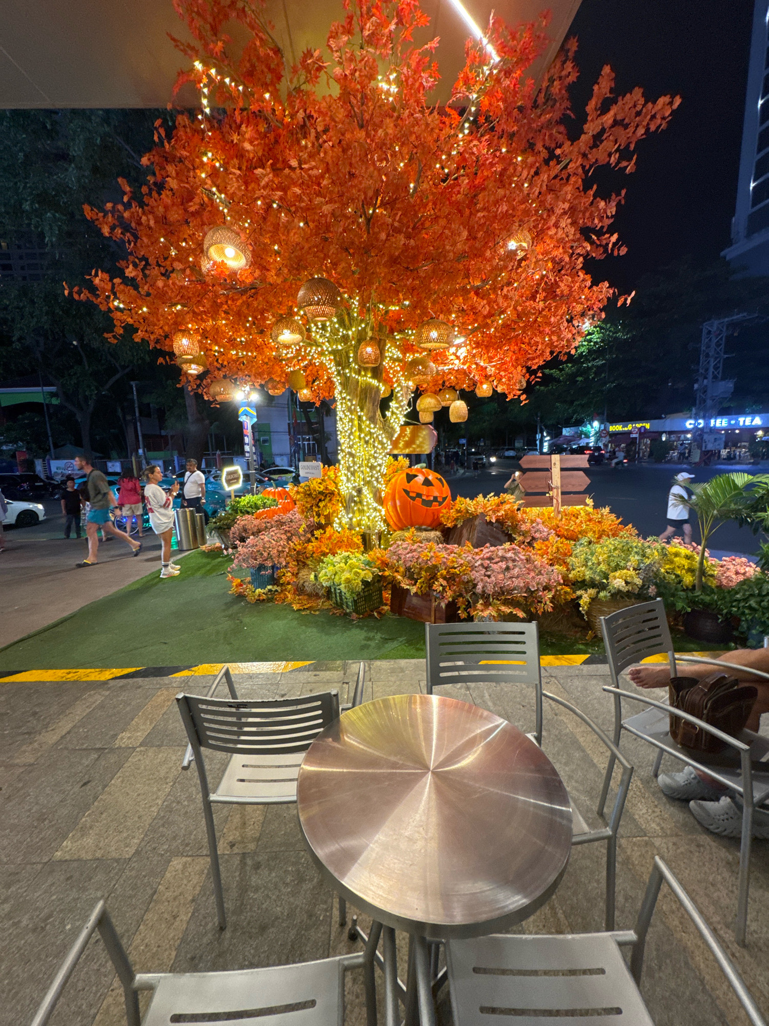 This is a nighttime street scene in Nha Trang, Vietnam, featuring a decorative setup for Halloween. In the foreground, a circular metal table is surrounded by four identical silver metal chairs, positioned on a patterned paved surface. Beyond the seating area, a bright green artificial turf covers a raised platform, demarcated by yellow and black hazard stripes. Dominating the background is an artificial tree adorned with vibrant orange leaves and numerous small white string lights, as well as woven lantern-like light fixtures. A large, carved jack-o'-lantern sits at the base of the tree, surrounded by an arrangement of colorful artificial flowers and foliage in shades of pink, orange, and yellow, interspersed with straw-like ground cover. A wooden directional sign with several blank arrows stands near the jack-o'-lantern.
To the left of the decoration, a few people are visible. A man wearing a light-colored t-shirt and blue shorts walks away from the camera. A woman in a white dress and athletic shoes stands facing the tree, holding a phone. Further back, another man in a white t-shirt and dark pants is visible. Cars and a motorbike are present on the street behind the decorative display, with shops and buildings lining the road. The overall ambiance suggests an outdoor seating area or walkway decorated for a seasonal event.