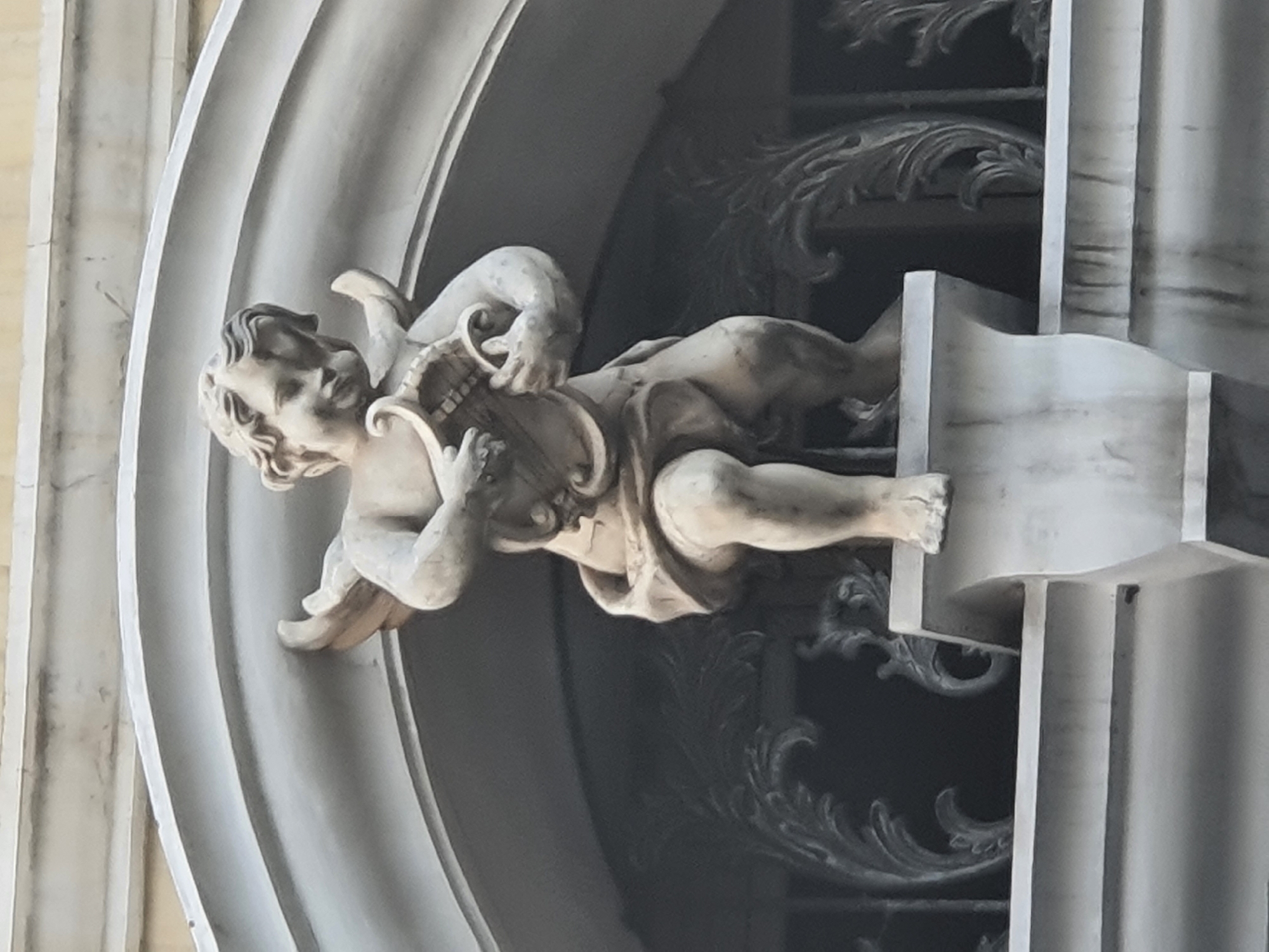 The image shows a stone sculpture of a cherub playing a lyre, positioned within an ornate architectural setting. The cherub, depicted with wings, is seated on a pedestal. The background features elaborate scrollwork and an arched structure. The sculpture appears to be made of light-colored stone, with visible weathering and some grime. The overall aesthetic suggests a classical or baroque architectural style. The image was taken in West Jakarta, Indonesia.