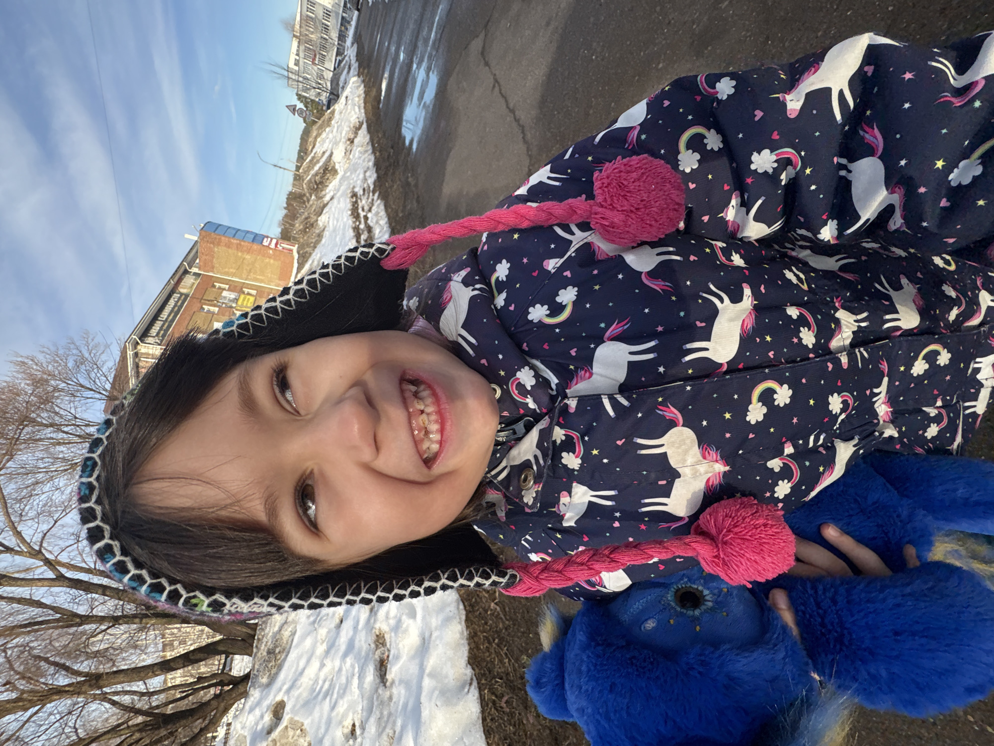 A young female child, estimated to be between 4 and 7 years old, is captured in a close-up outdoor shot, positioned near Lenin Street, 30, in Chaikovsky, Russia. She wears a black winter hat with a multi-colored, honeycomb-patterned brim and two bright pink braided tassels that terminate in pom-poms. Her dark brown hair is visible. The child is dressed in a dark blue padded winter jacket patterned with white unicorns, rainbows, and stars. She displays a wide, open-mouthed smile, looking upwards and to the right, revealing primary teeth, some of which show visible decay or wear. In her left hand, she holds a large, electric blue plush toy featuring distinct blue eyes and an intricate facial design.
The immediate ground appears to be dark asphalt with scattered puddles and patches of melting snow. To the child's left and behind her, snow covers an incline where bare deciduous trees stand. The background features urban structures under a bright blue sky with sparse clouds. Prominently in the mid-ground is a multi-story building constructed of yellow-brown brick with white-framed windows. A large yellow banner affixed to its side clearly displays Russian text, reading "ДЕТСКИЙ МИР" (Children's World) and "МЫ ОТКРЫЛИСЬ" (We are open). Further to the right in the background, another lighter-colored, multi-story building with numerous windows and a circular road sign with a red border are visible. The scene suggests a clear late winter or early spring day.