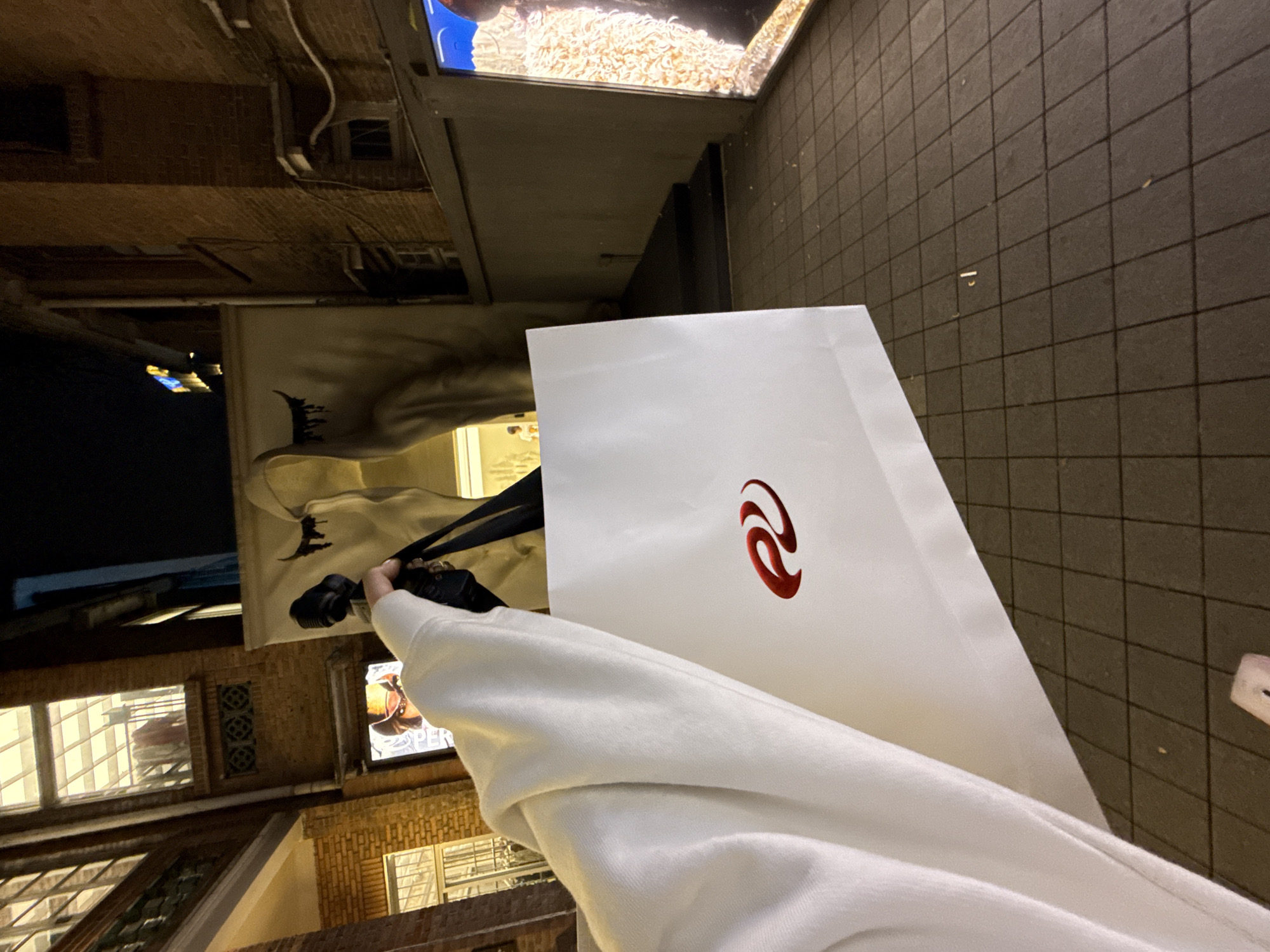 A person wearing a white sweatshirt holds a white shopping bag with a red logo on a tiled sidewalk in Tianhe District, China. In the background are buildings with illuminated windows and signage. The viewpoint is from the person holding the bag.