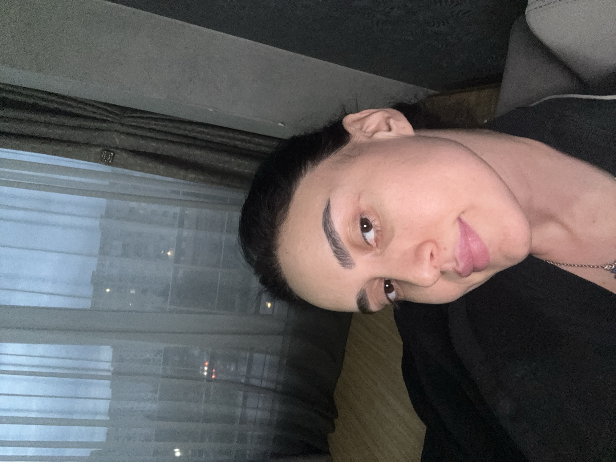 A rotated close-up portrait shows an individual, likely female, looking towards the viewer indoors in Tbilisi, Georgia. She has dark hair pulled back, distinct dark eyebrows, and light brown eyes. Her expression is a subtle, somewhat playful smile, with her tongue slightly visible. She is wearing a dark top and a delicate silver necklace with a pendant.
The background reveals a window on the left, partially obscured by blinds or sheer curtains. Through the window, an urban environment at dusk or night is visible, with distant building lights and reflections on the glass suggesting a city scene. A dark wall or ceiling section is in the top left, and a dark, possibly wooden, surface is in the bottom right of the frame. The overall lighting is dim, indicating an evening setting. There is no visible text in the image.