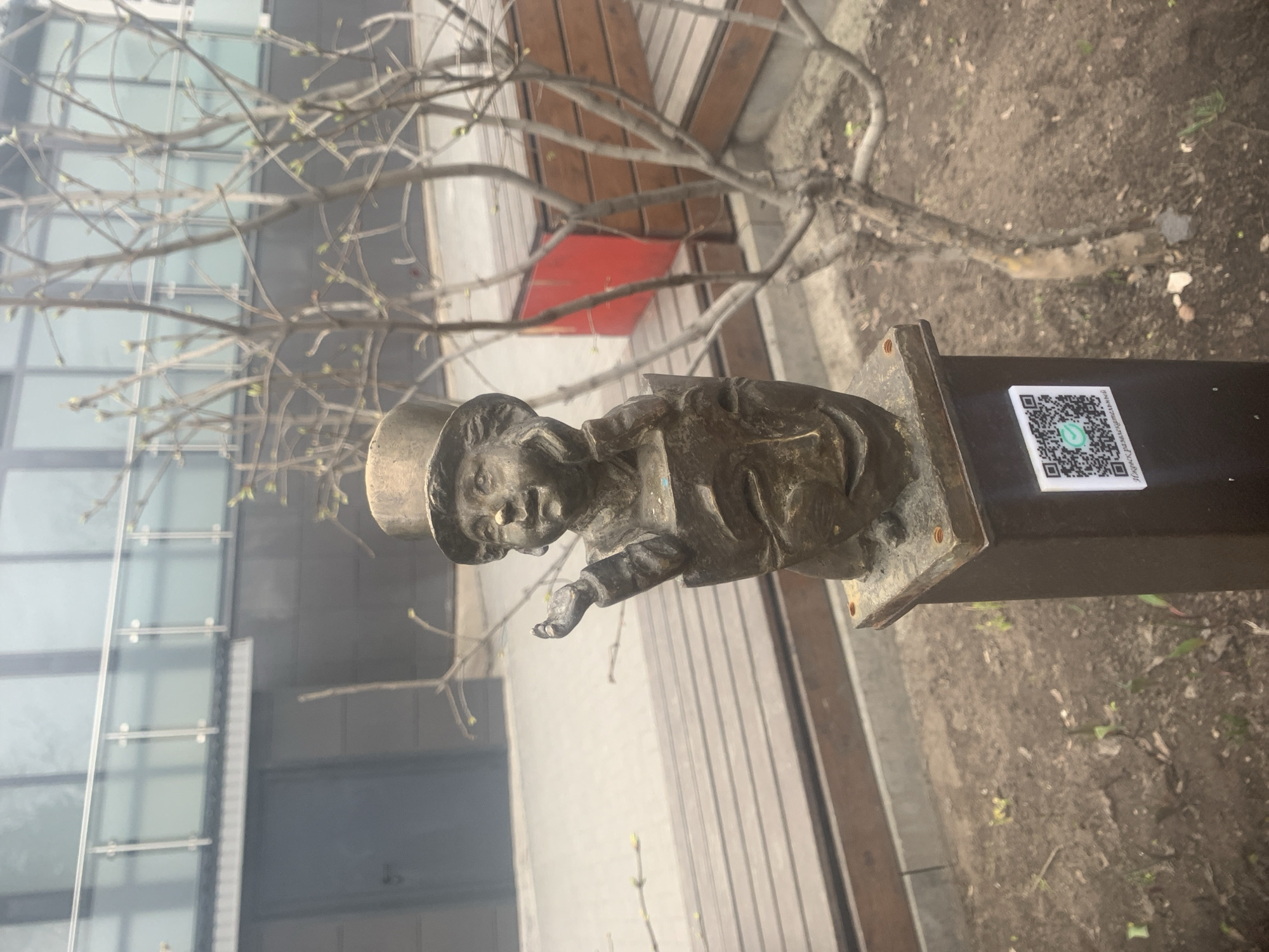 A bronze statue of a small, uniformed person with a hat and outstretched hand stands on a dark metal post in Izhevsk, Russia. A QR code sticker is adhered to the side of the post. Behind the statue is a light gray wooden plank walkway. Above and to the right of the statue is a tree branch. Behind the tree and statue is a glass-paneled building. To the right of the walkway is a dirt-covered area.