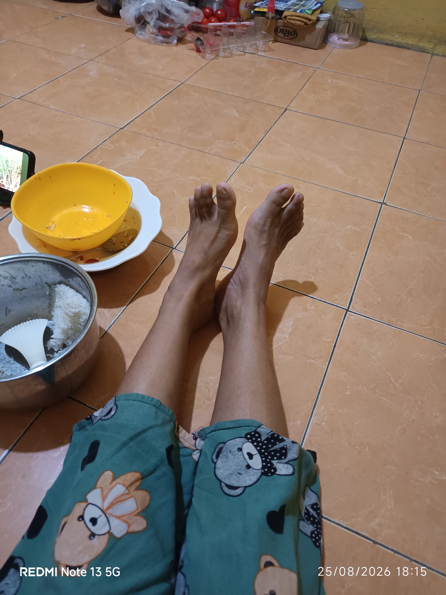 The image shows a pair of legs and feet resting on a tiled floor. The person is wearing green pajama bottoms with a teddy bear pattern. To the left, a yellow bowl with some food residue and a spoon is placed on a white plate. Next to it, a metal pot contains white rice and a white food scoop. A cell phone is visible on the left side of the frame, displaying a green image. In the background, there are some items on a shelf, including food packaging and a clear plastic cup. The floor is tiled in an orange-brown color. The timestamp in the lower right corner indicates the date 25/08/2026 and time 18:15. The text "REDMI Note 13 5G" is present in the lower left corner. The scene appears to be an indoor domestic setting, possibly a kitchen or dining area. Given the information, it is not possible to determine the specific location in the city of Talaga Sari, Indonesia.