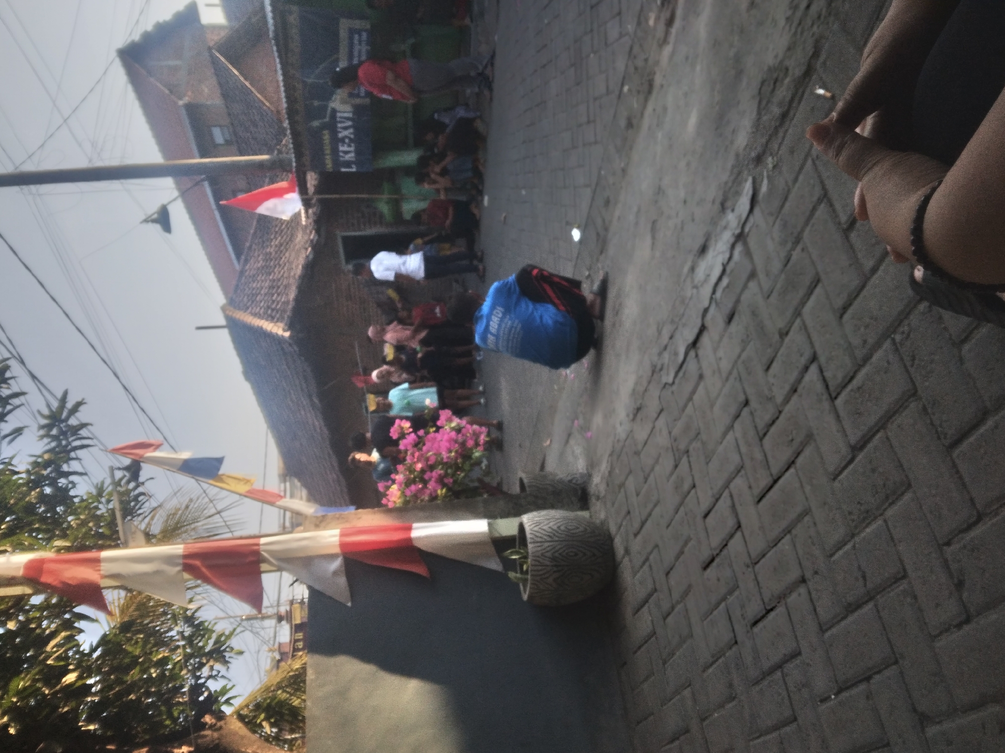In Wringinanom, Indonesia, a street scene unfolds with a focus on a young person in a blue shirt squatting in the foreground. The individual's back is to the camera, and they are wearing red and black striped pants. Behind them, a group of people are gathered, some standing and some sitting on the ground. A building with a red-tiled roof is visible in the background, with a banner displaying text that appears to be in Indonesian. Flags, including red and white Indonesian flags, are displayed on poles, some with red and white stripes, and one with red, white, and blue. There are trees and a potted plant with pink flowers in the scene, and the ground is paved with interlocking bricks. The overall atmosphere suggests a community gathering or event.