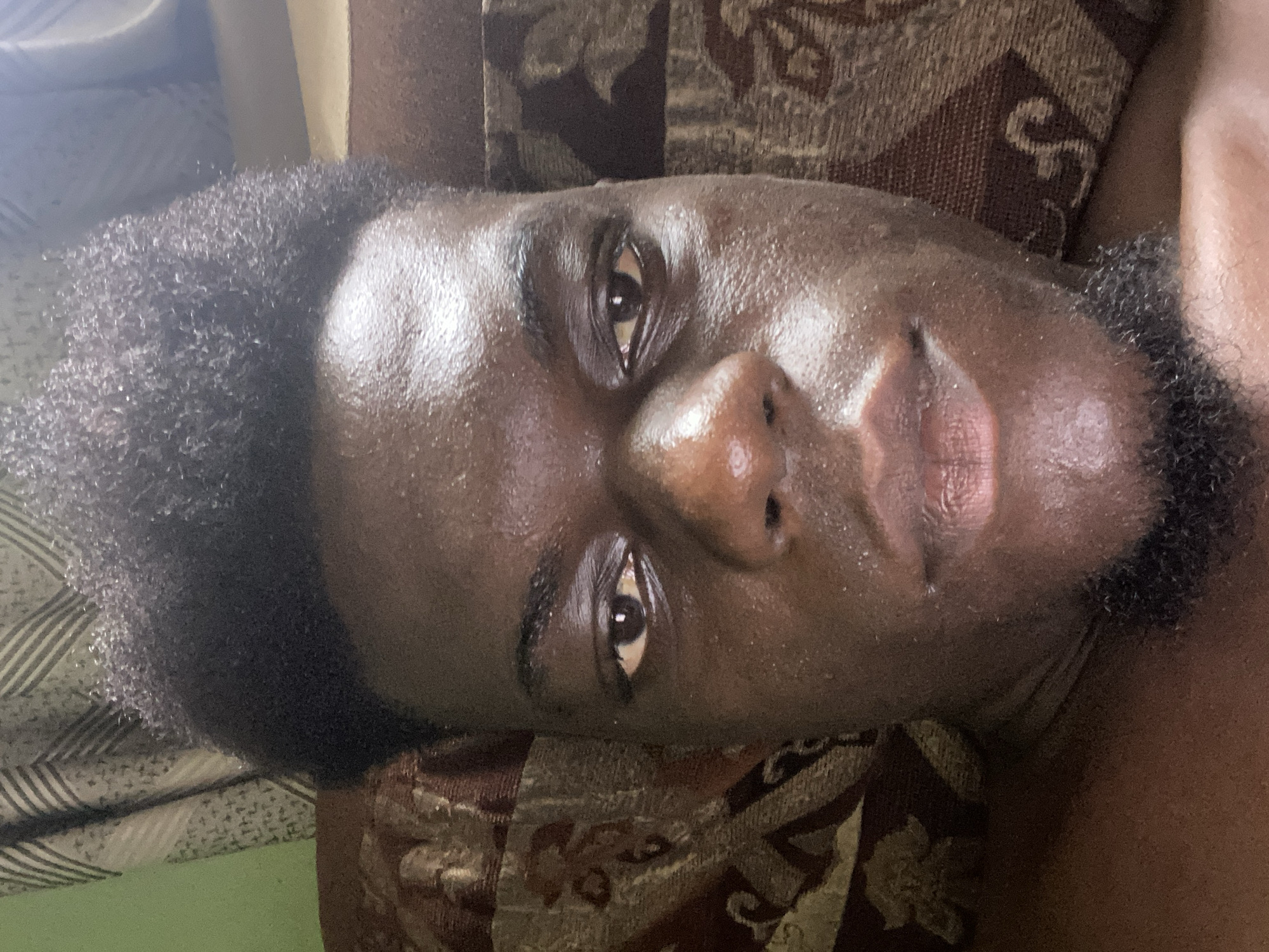 A close-up portrait shows a person with dark skin and tightly coiled dark hair, lying down indoors. The person's head is tilted to the left, with their gaze directed towards the camera, conveying a neutral and calm expression. Their skin has a noticeable sheen or oily appearance, particularly on the forehead, nose, and cheeks, where highlights are prominent. A short, dark beard or stubble is visible on their chin and jawline. Their eyes are dark and appear slightly narrow or partially closed, revealing the dark irises. The lips are full.
The background suggests an indoor setting, likely a sofa or bed, covered with various textiles. Prominently featured is a patterned fabric in shades of brown, gold, and cream, adorned with intricate, possibly ornamental, designs. Other background elements include a striped fabric and a solid green area, adding to the layered appearance of the setting. The lighting appears to be artificial indoor light. No specific activity is depicted other than posing for the portrait. There is no visible text in the image.