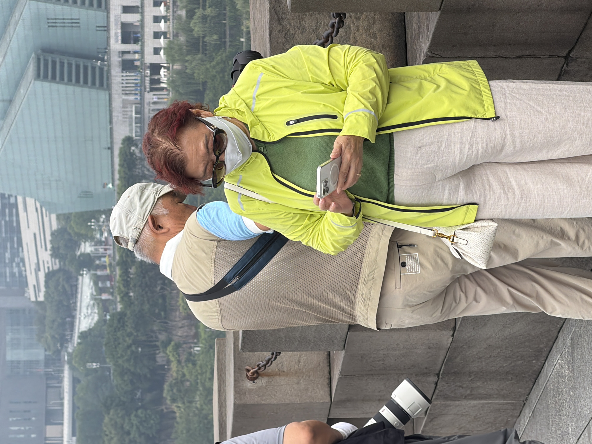 In the Yuzhong District of China, two individuals stand on a stone structure. A person wearing a tan vest over a blue shirt and a light-colored cap is viewed from the back, with their head tilted down. To their right, a woman in a bright yellow jacket and light-colored pants is looking at her smartphone, wearing sunglasses and a white mask. A small white handbag hangs from her left shoulder. In the background, buildings and trees are visible. Part of a camera lens is visible on the left side of the frame. The stone structure appears to be part of a bridge or walkway.