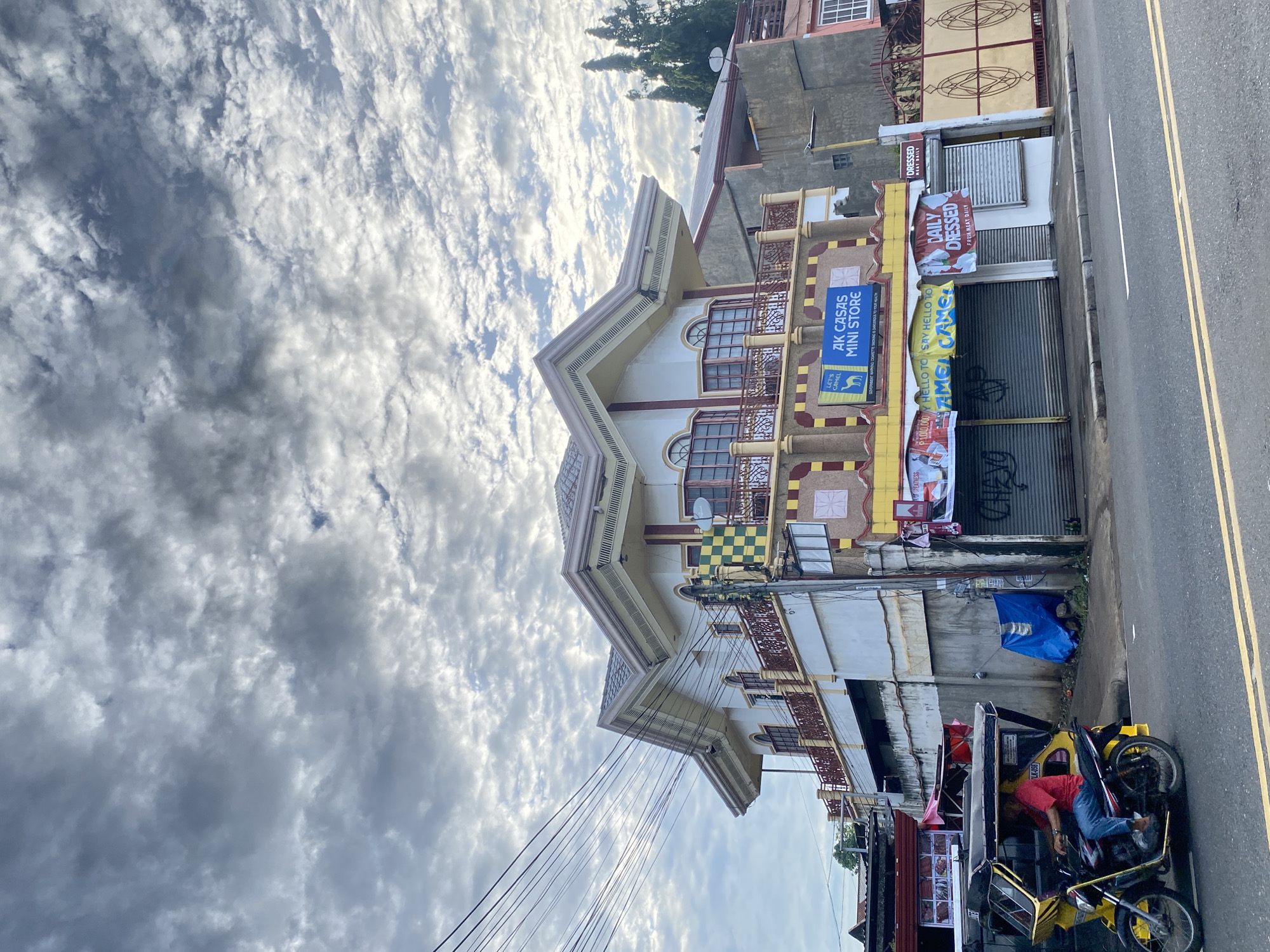 The image captures a street-level view of a two-story building in Davao City, Philippines, under a cloudy sky. The building features a prominent roof with decorative eaves and balconies with railings. The ground floor houses a commercial establishment with a sign that reads "AK CASAS MINI STORE" and another banner advertising "DAILY DRESSED" and "FRESH MEAT DAILY". A tri-cycle, a common mode of transport in the Philippines, is visible on the street with a person wearing a red shirt and blue jeans driving it. The street is paved with asphalt and has yellow lane markings. Electrical wires are strung across the top of the scene. The surrounding buildings are a mix of concrete structures, some with decorative elements and metal gates. The overall impression is of a busy urban commercial area.