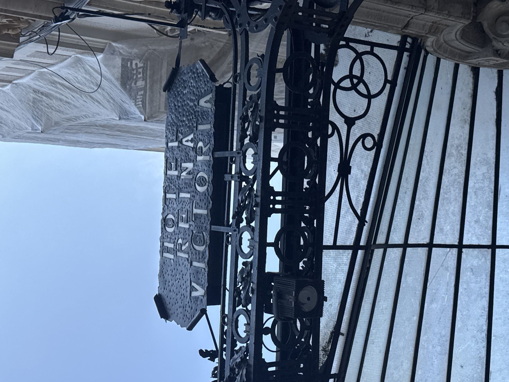 The image captures a close-up view of a dark, ornate metal sign that reads "HOTEL REINA VICTORIA" against a bright, overcast sky. The sign is attached to a wrought-iron structure that appears to be an entrance canopy, with intricate scrollwork and circular decorative elements. Below the sign, a glass-paneled roof is visible, supported by dark metal beams. To the right of the sign, a building facade is partially obscured by white sheeting, suggesting ongoing construction or renovation. The architectural style of the building and the wrought-iron work suggest a European setting, possibly historical. Without further information or visual cues such as street signs or distinctive landmarks, pinpointing the exact location within Valencia, Spain, is not possible based solely on this image. However, the "Hotel Reina Victoria" is a known landmark in Valencia, located at Calle Correos, 8, near the Plaza del Ayuntamiento.