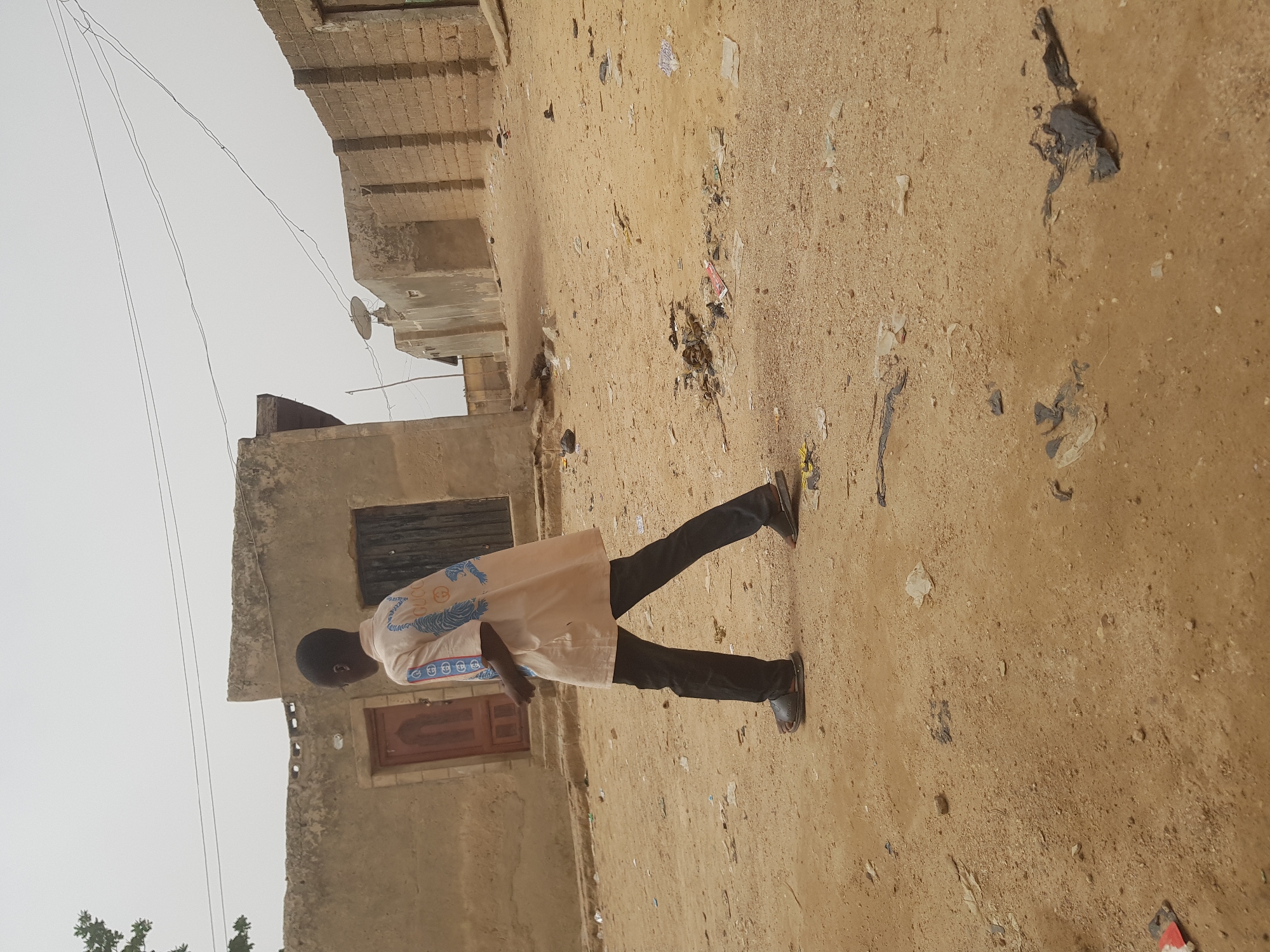 A medium shot of a young Black male is seen in Dutsinma B, Nigeria. He is in the foreground of the image, walking away, wearing dark pants, slides, and a light tan long-sleeved shirt with embroidered detail around the chest. To the left of the subject is a tan building with a brown door and dark windows. To the right of the subject is a dirt ground filled with litter. Above the building on the left is a cloudy, pale sky with electrical wires, and a satellite dish.