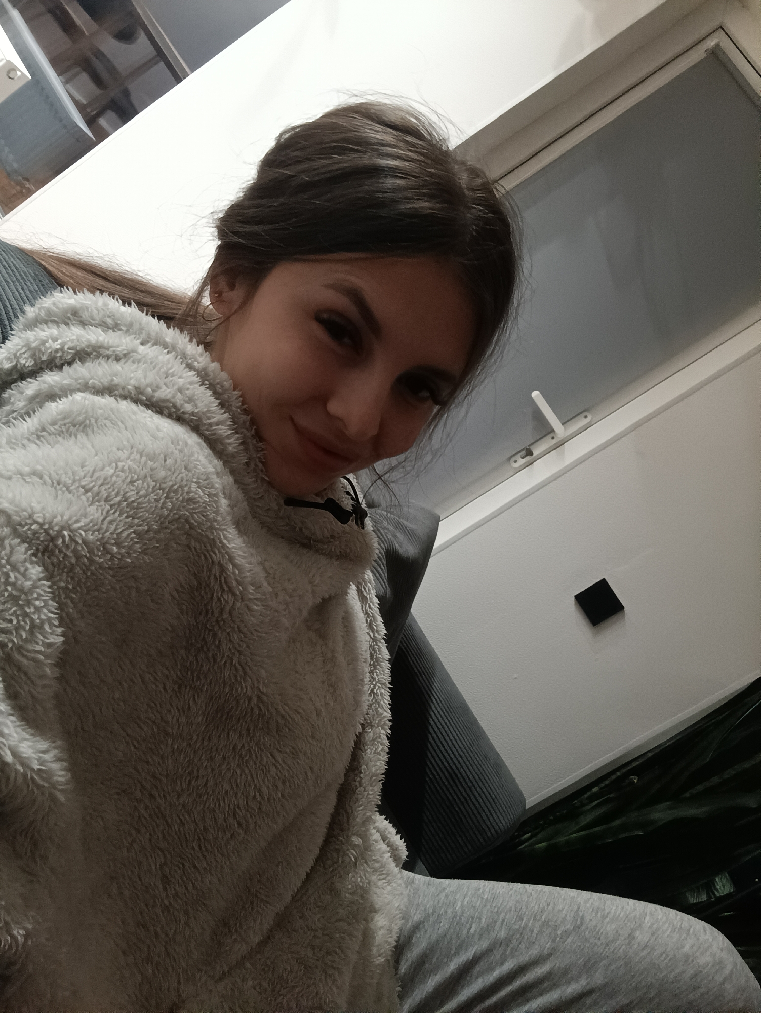 A selfie photograph shows a woman indoors. She is wearing a fuzzy gray sweater and gray pants, suggesting leisurewear. She has dark hair pulled back, and is smiling subtly. Behind her is a window with a white frame and a white handle. The wall is white and has a small, square, black object affixed to it. The seat she is sitting on appears to be gray. There are plants to her right. There is no information to determine the location of the photograph is Сепыч, Russia.