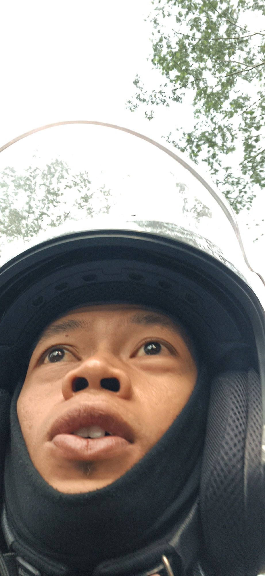 A low-angle, close-up selfie shows a person wearing a black helmet and black neck gaiter, looking upwards. The person's face, from the nose to the chin, is prominently featured. The helmet has a reflective visor that shows a blurred image of green foliage against a bright sky. The background is predominantly a bright, overcast sky with scattered green leaves, suggesting an outdoor environment, possibly a street in Mlobo, Indonesia, given the context. The person's eyes are open and directed upwards, and their mouth is slightly open. The focus is sharp on the person's face, while the helmet and background are slightly out of focus.