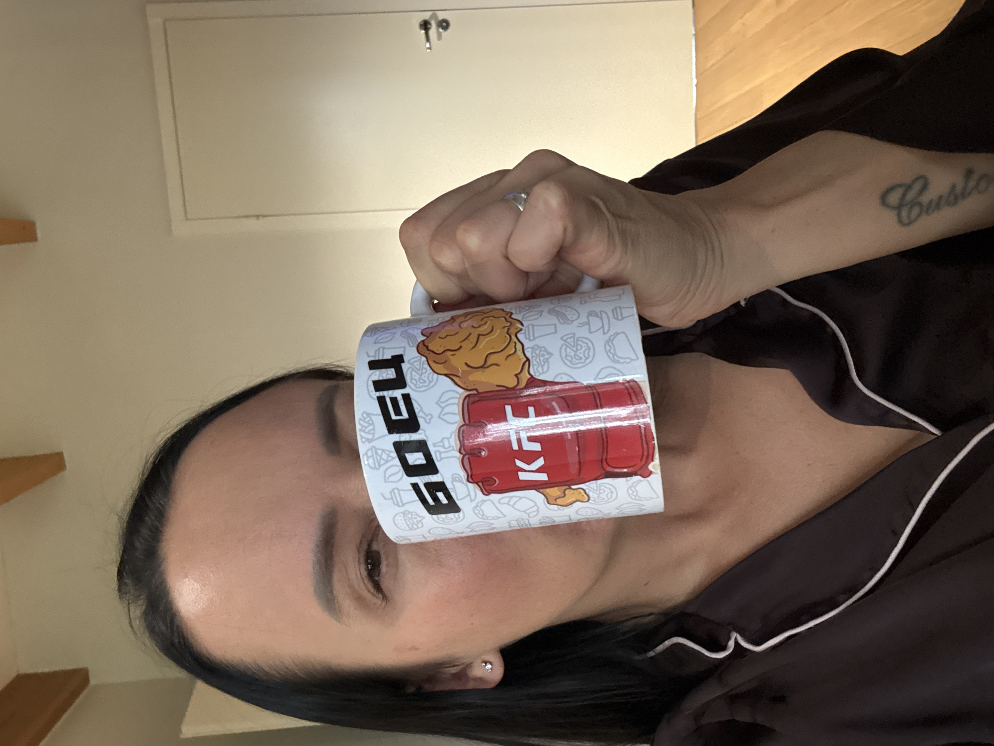 A woman is holding a mug in front of her face, obscuring her eyes and forehead. The mug is white with a colorful cartoon design featuring fried chicken and the text "GOEU" and "KF". She appears to be indoors, possibly in a home, as indicated by the wall and wooden floor visible in the background. The woman is wearing a dark, silky robe with white trim. A tattoo that reads "Custo" is visible on her forearm. The lighting suggests it might be daytime.
