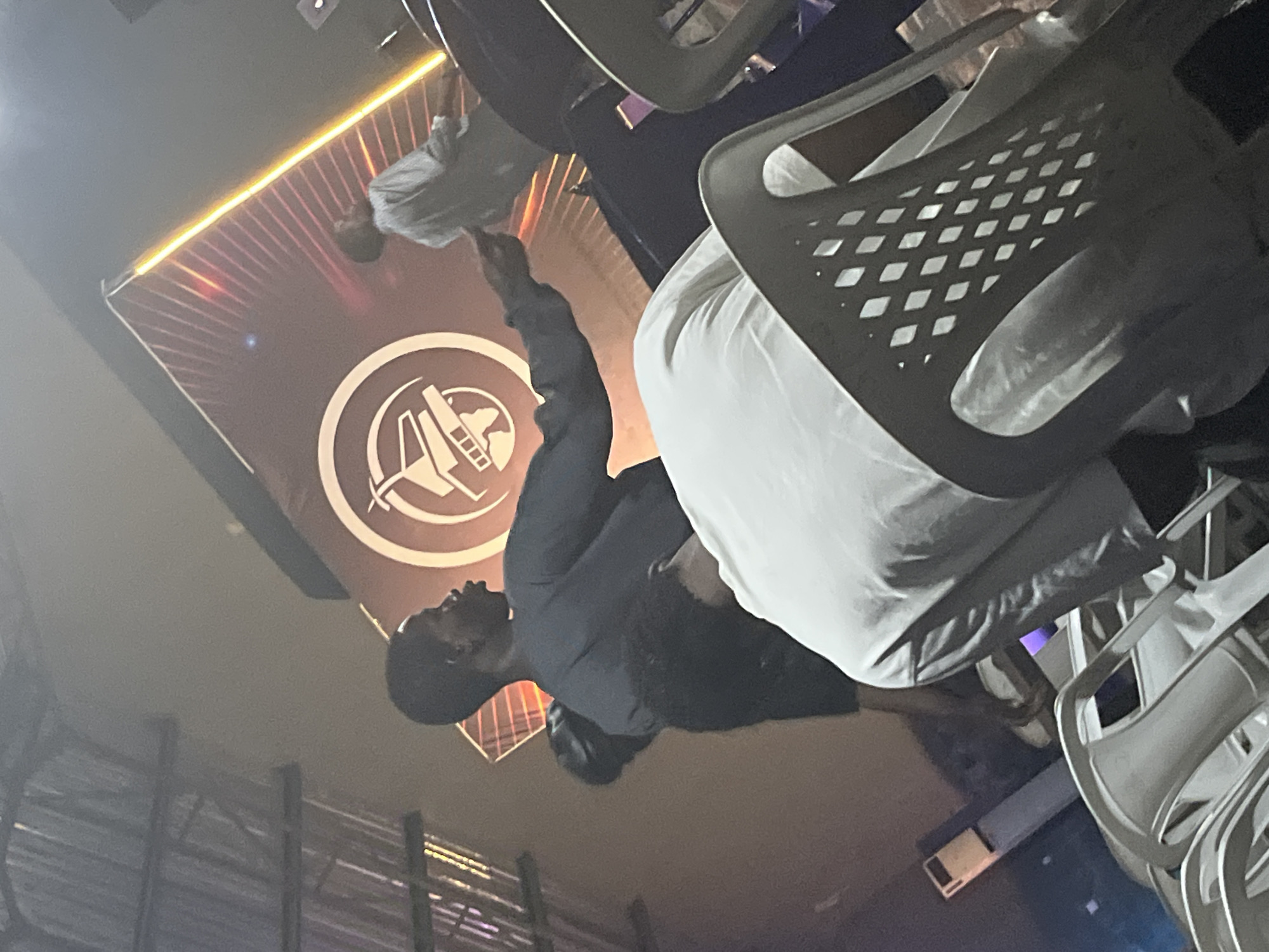 The image depicts an indoor scene, possibly a hall or venue, with a stage area in the background. A large, circular logo with an airplane and world map graphic is prominently displayed on a banner, illuminated by orange light that emanates from the sides. In the foreground, several white plastic chairs are arranged, some occupied by people.
A young Black man wearing a dark long-sleeved shirt is standing and appears to be interacting with someone off-camera, extending his left arm forward with his hand open. Behind him, another person, also appearing to be a young Black man, is seated and leaning forward, with their head down. A third person, wearing a light-colored shirt, is partially visible further back, near the stage. The ceiling appears to be made of corrugated metal or similar material, suggesting a functional rather than a formal setting. The lighting is dramatic, with strong contrasts between illuminated areas and shadows. The overall ambiance suggests an event or gathering is taking place.
