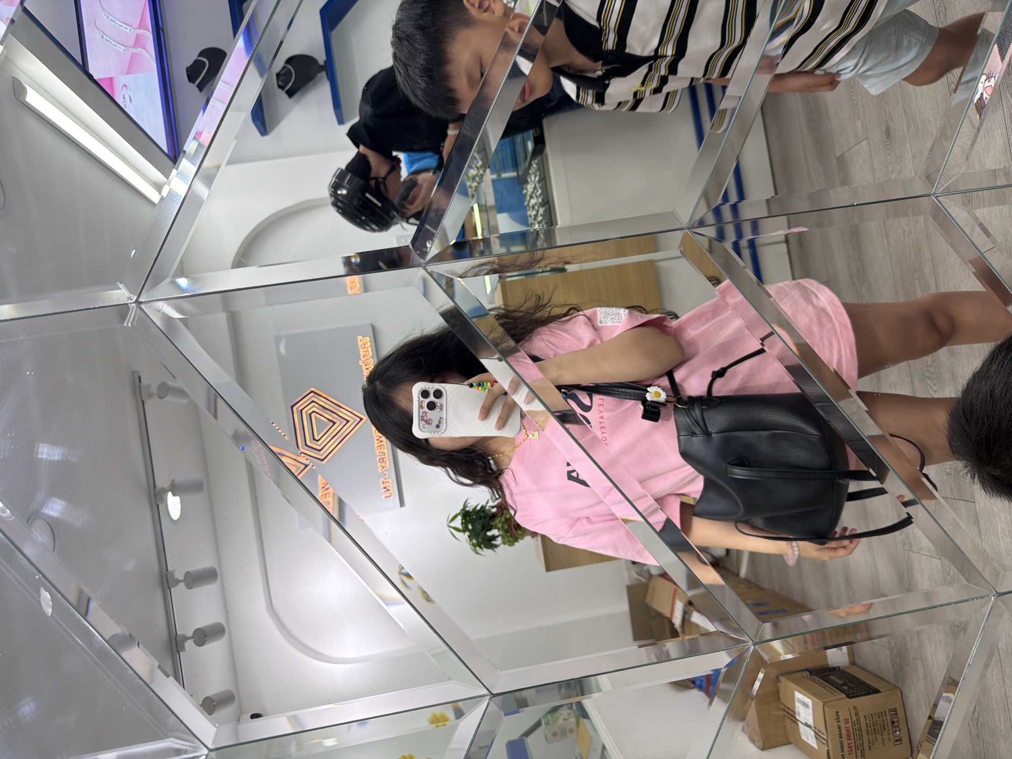 The image captures an indoor scene within a modern, highly reflective space, likely a shop or exhibition, characterized by numerous mirrors arranged in geometric patterns on the walls. The original image is rotated counter-clockwise, showing a kaleidoscopic effect with fragmented reflections.
In the foreground, a young woman with long dark hair, dressed in a pink t-shirt and light-colored shorts, is taking a selfie with a white smartphone. The phone has a distinctive patterned case and a colorful beaded strap. She carries a black shoulder bag, which features a white daisy clip. Her reflection reveals a gold necklace and a light-colored bracelet on her wrist.
Multiple reflections populate the mirrored panels, showcasing other individuals and parts of the room. A young boy with dark hair, wearing a black and white striped shirt with yellow accents and light blue shorts, is visible in an upper reflection. Another person, possibly an adult, wearing a dark top and a black protective helmet or headgear, is also reflected in the upper part of the image.
The background elements, fractured by the mirror arrangement, include white walls, bright strip lighting, and what appear to be display cases or shelves, some showing jewelry or other items. A digital screen displaying jewelry designs is visible in one reflection. The floor is a light-colored wood or laminate, and several cardboard boxes are seen in the lower reflections.
A notable detail is an orange geometric logo, resembling a nested diamond shape, visible on a mirrored surface, accompanied by mirrored text that is difficult to decipher but appears to be related to jewelry, possibly "DIAMOND JEWELRY" or a similar phrase in a language other than English, given the location. The environment is brightly lit, indicating indoor artificial lighting. This dynamic scene takes place in Đà Nẵng, Vietnam.