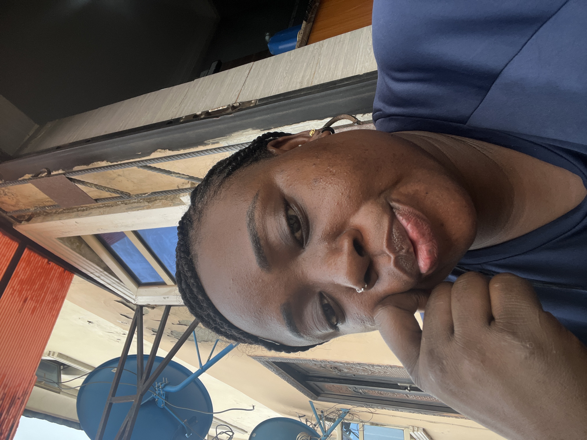 This vertical selfie features a dark-skinned woman looking directly at the camera with a subtle, pleasant expression. She has dark, braided hair and wears a dark blue top. Notable accessories include a small silver nose ring and a delicate gold earring on her right ear. Her right hand is positioned near her chin and mouth.
The background suggests an outdoor, possibly urban or semi-urban setting in Dushepe, Nigeria. Various building structures are visible, including a light-colored facade with dark ledges and what appears to be exposed rebar in parts. A window with blue-tinted glass is also seen in the upper left quadrant. Towards the lower left, multiple blue satellite dishes and associated metal support structures are clearly visible. A section of an orange, corrugated surface is present on the far left. The bright, even lighting indicates it is daytime, likely under sunny conditions. There is no visible text in the image.