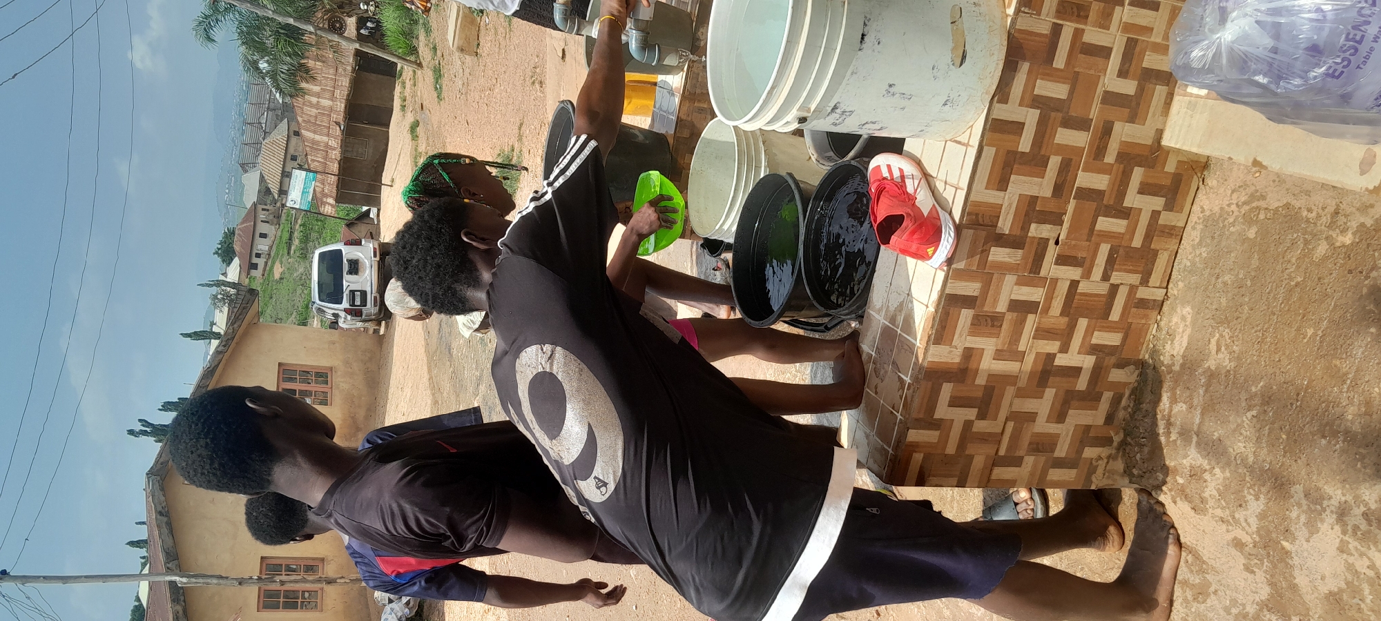 In Abuja, Nigeria, a group of young individuals are gathered around an outdoor water source. The scene unfolds in a dusty, outdoor environment, possibly a community water point or a private residence. A white SUV is parked in the background, suggesting it might belong to someone in the vicinity.
The foreground is dominated by several large buckets, some filled with water. One person, wearing a black t-shirt with a large white "9" on the back, is leaning over to interact with the water. Another young person, with distinctive green braids, is also looking towards the water. A bright red and white sneaker rests on the tiled surface next to the buckets.
The tiled surface appears to be part of a counter or washing area. Beyond the immediate scene, the environment shows a collection of buildings with corrugated iron roofs, typical of residential areas in Nigeria. The overall impression is one of everyday life, with people accessing a vital resource. The ground is a mix of compacted dirt and scattered debris. The sky is visible overhead, with some clouds present.