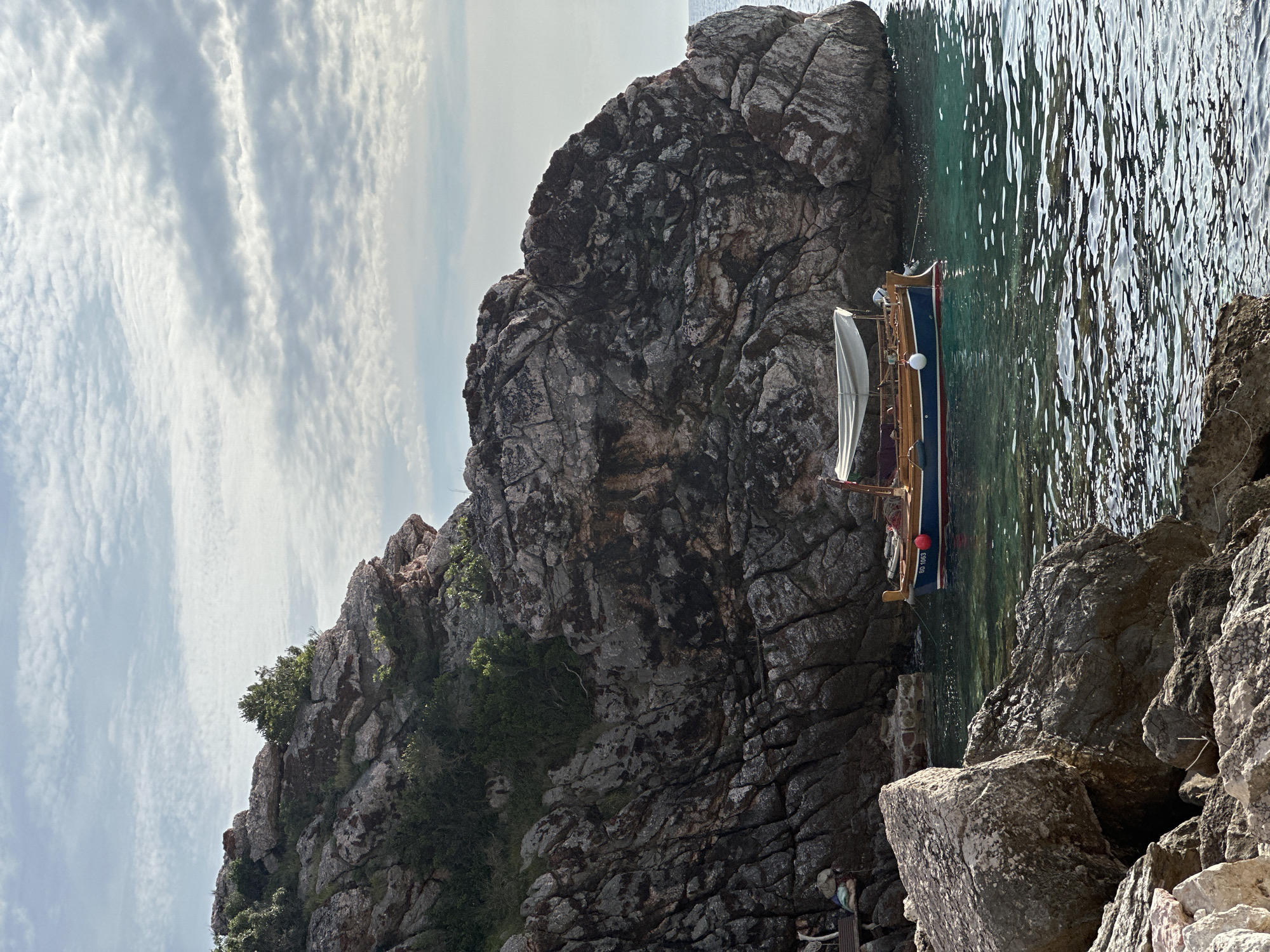 This image depicts a coastal scene in Rijeka Reževići, Montenegro. The dominant features are large, rugged rock formations that rise steeply from the water, occupying the left and central-right portions of the frame. These rocks are dark grey and brownish, displaying extensive fracturing and weathering, with patches of green scrub and small trees growing in crevices. The sky above is light blue with scattered, wispy stratocumulus clouds.
In the clear, turquoise-hued water, a small, traditional wooden boat is moored near the base of the rock face. The boat has a blue hull, a red waterline stripe, and natural wood upper trim. A light-colored canvas canopy is deployed over its midsection. A white spherical fender is visible on the port side of the boat, and the registration number "BD 1063" is clearly legible on its stern.
To the left of the boat, a narrow indentation in the rock face accommodates a dark wooden bench or platform. On or near this platform, several light-colored bundles or bags are visible, some partially covered with what appears to be blue or green material, along with ropes or lines. The foreground comprises large, irregular rock fragments, indicating the viewpoint is from a rocky shore. The water's surface shows gentle ripples, reflecting the sky and light conditions. There are no individuals visible in the scene.
