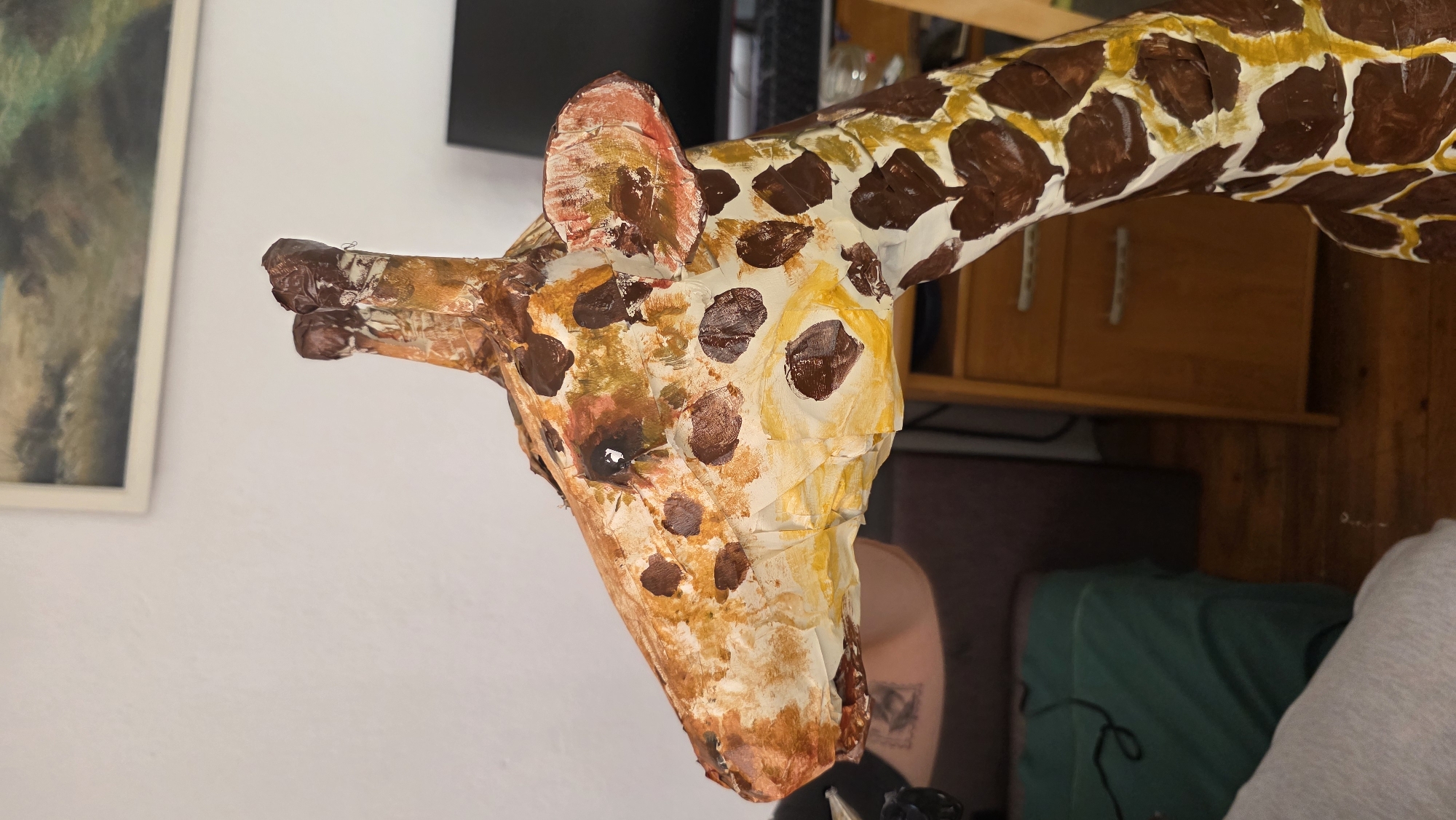 A close-up shot of a handmade giraffe head sculpture, with the head and neck featuring tan and white sections with brown spots. The giraffe has small ears, horns, and visible facial features. The background includes a white wall, a framed picture, a computer monitor, wooden cabinets, and a green object. The location in the city of Blagoevgrad, Bulgaria is not evident in the image.