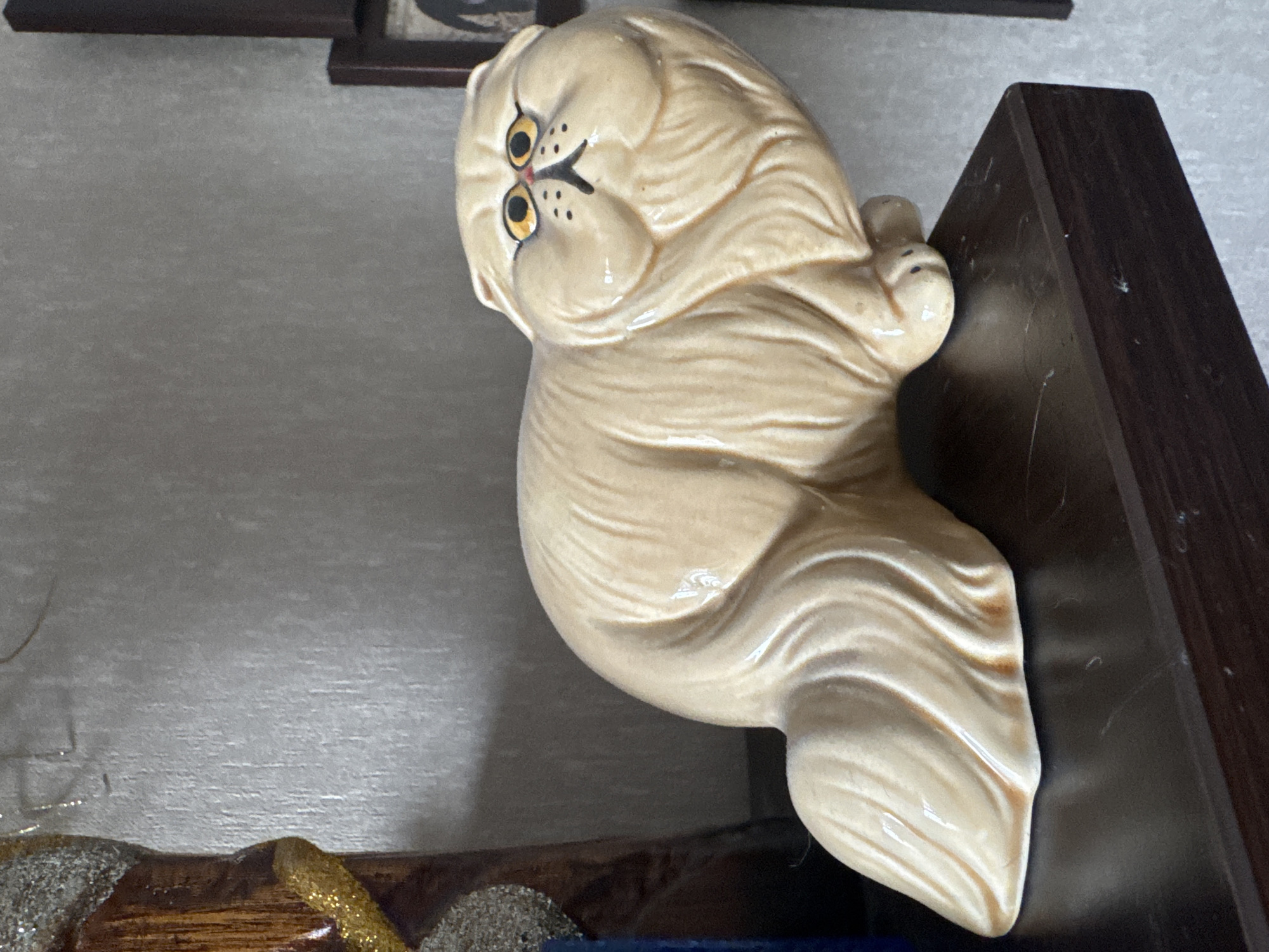 A tan ceramic cat figurine with large yellow eyes and black features sits on a dark brown shelf. The figurine is positioned on the right side of the frame, facing left. The background is a textured gray wall. To the left of the figurine, a portion of a wooden object with glittery embellishments is visible. The shelf appears to be within an indoor setting. The specific location in Bishkek City, Kyrgyzstan is not discernible from the image.