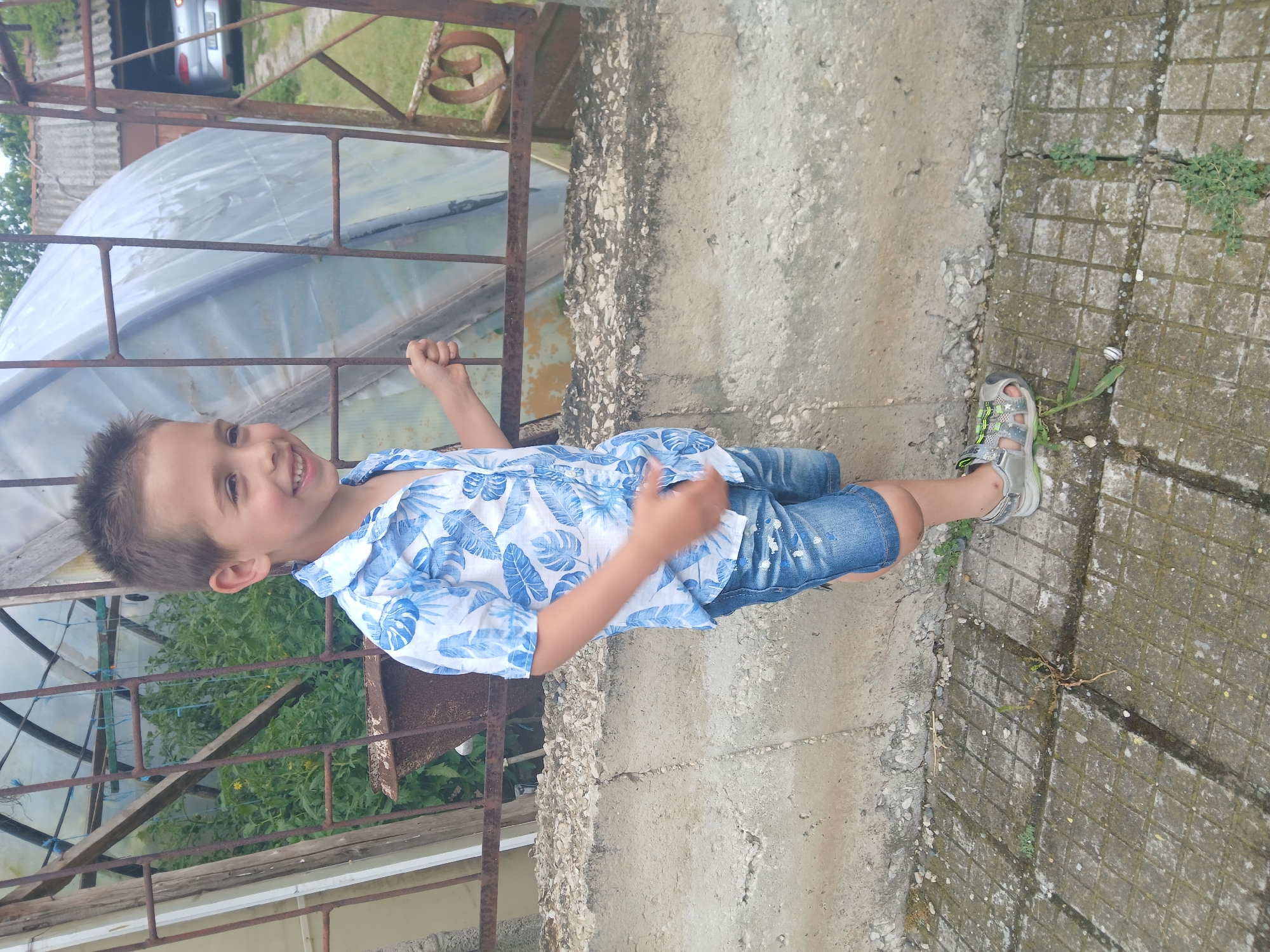 A young boy with brown hair is smiling and looking to his right. He is wearing a blue and white floral short-sleeved shirt, denim shorts with white paint splatters, and silver sandals. He is standing on a tiled surface in front of a concrete wall. Behind him, a rusty metal fence encloses a greenhouse filled with green plants. In the background, a car is visible parked near a building with a grey roof. The scene appears to be outdoors, possibly in a rural or suburban area.