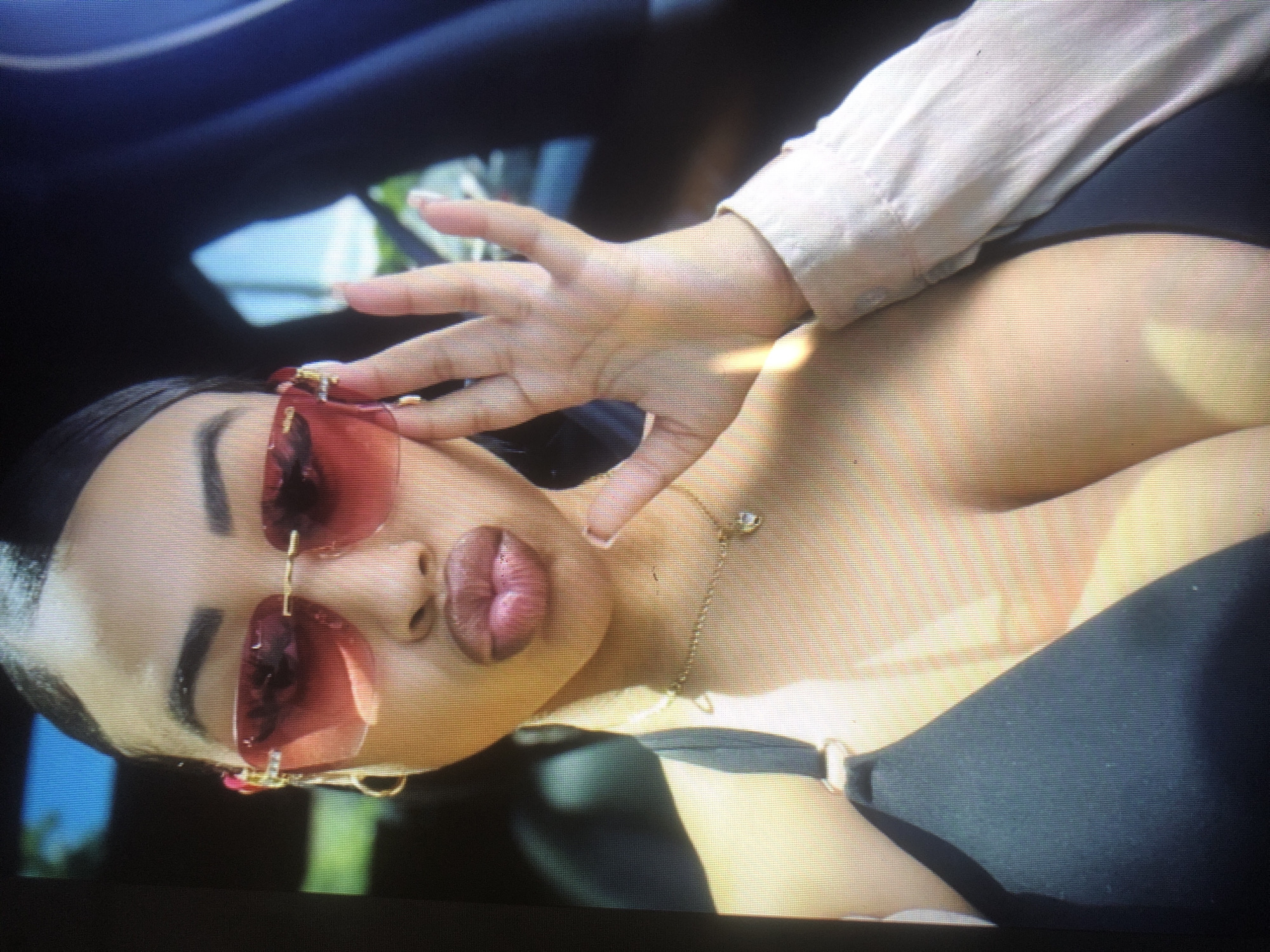 The image shows a close-up of a young woman's face and upper body, angled from the side. She is wearing large, red-tinted sunglasses and has her hand raised near her face. Her lips are full and appear to be painted with a matte lipstick. A delicate gold chain necklace with a small pendant rests on her collarbone. She is wearing a light-colored, possibly silk, blouse, and a dark garment that appears to be a strapless top or bra. The lighting suggests it is daytime, with natural light illuminating her skin. The background is dark and out of focus, with some hints of blue and green, possibly indicating an outdoor setting or the interior of a vehicle. The overall mood is suggestive and alluring, with the woman's gaze directed away from the camera, partially obscured by her sunglasses. There is no visible text in the image.
