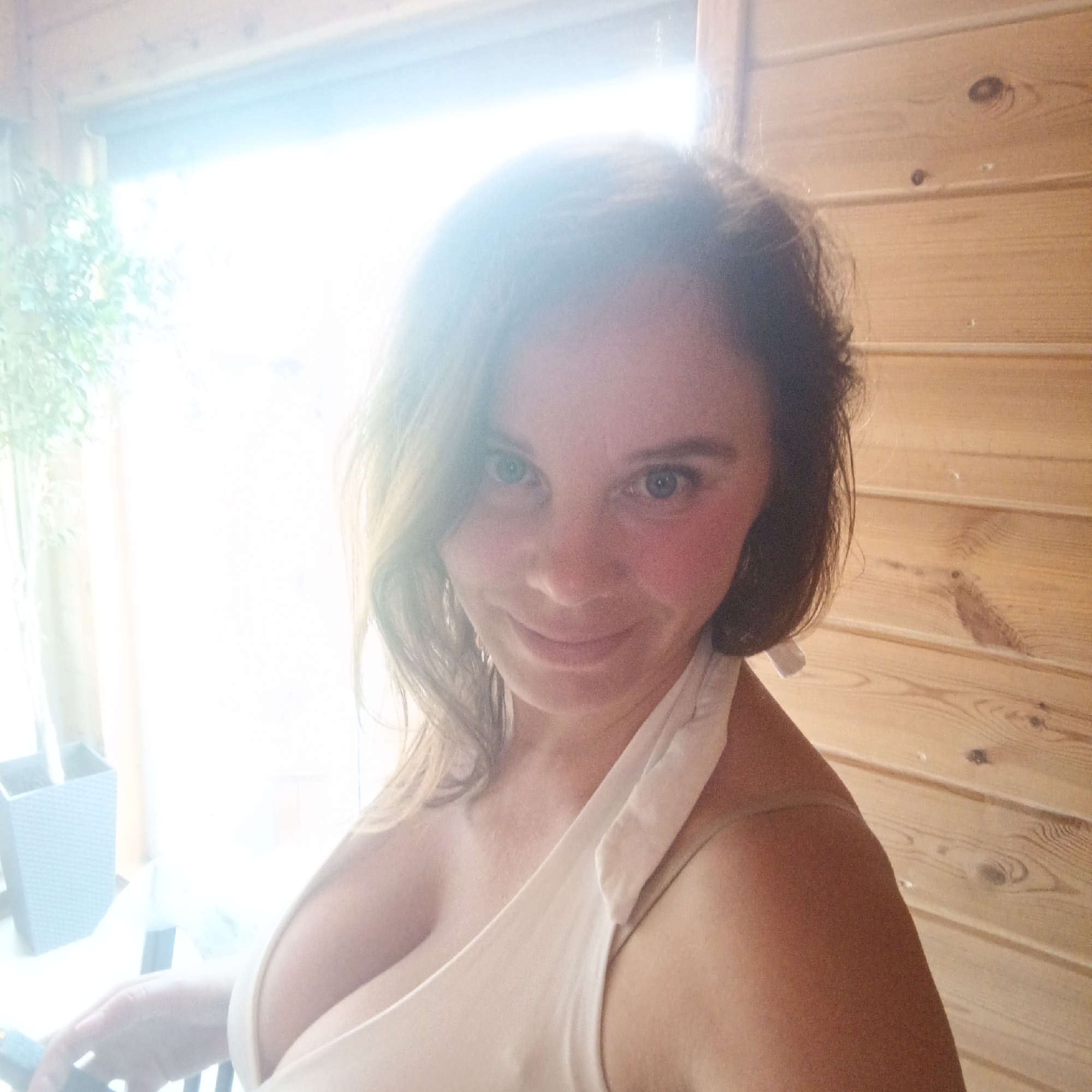 A woman with brown hair and blue eyes is pictured in a close-up selfie. She is wearing a white tank top. The background consists of wooden walls and a brightly lit window. The image is taken indoors, likely in a residential setting. There are no clear indicators of the specific location within Chaikovsky, Russia, beyond it being an indoor scene.