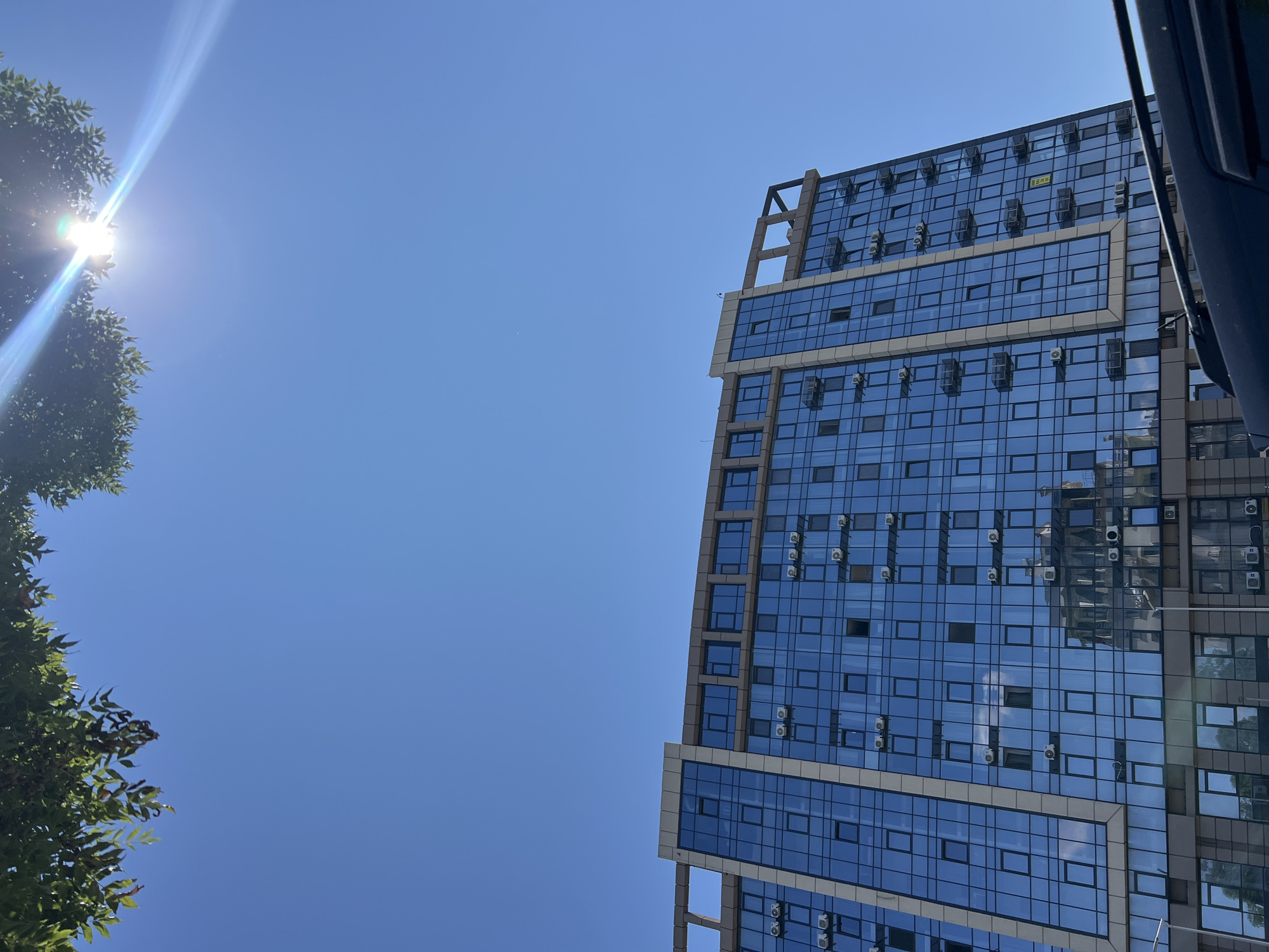 In Fontanka, Ukraine, a modern, multi-story building with a blue glass facade dominates the lower portion of the image under a clear, bright blue sky. The building features numerous windows and air conditioning units mounted on the exterior. Sunlight glints off the glass, and reflections of other buildings and the sky are visible. Green tree foliage hangs into the frame from the top left corner, partially obscuring a lens flare from the sun. A dark-colored vehicle is partially visible in the foreground at the bottom.
