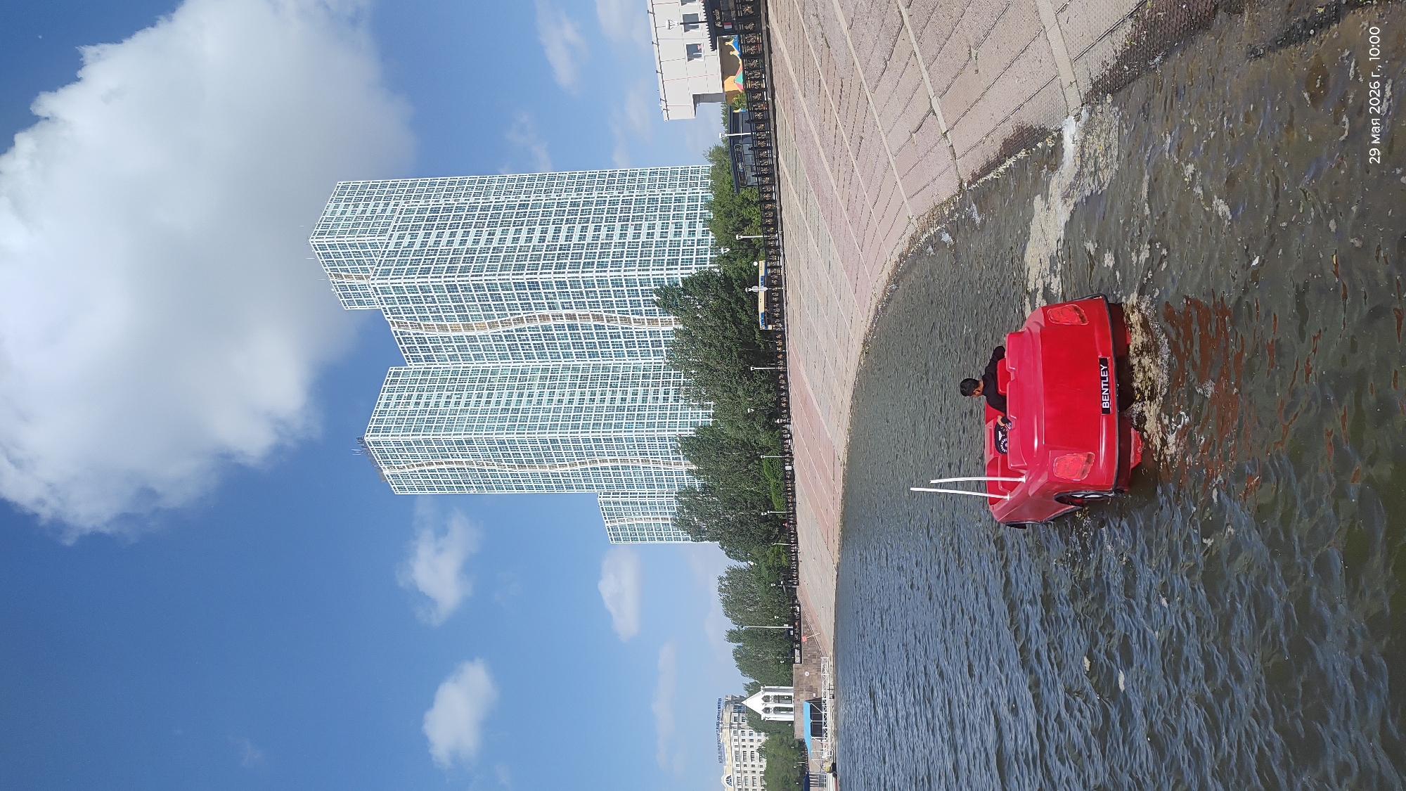 A landscape-oriented shot shows a red pedal boat on a body of water near a brick-paved embankment with a child in the boat. The boat has "BENTLEY" written on the stern and is close to the embankment. Two skyscrapers with a grid-like pattern dominate the background. The buildings are separated by trees, and other buildings can be seen in the distance. The sky is blue with scattered white clouds. The date "29 May 2026 г., 10:00" is imprinted on the top right corner of the image. The location appears to be a body of water in Astana, Kazakhstan, with the skyscrapers potentially located along the Ishim River.