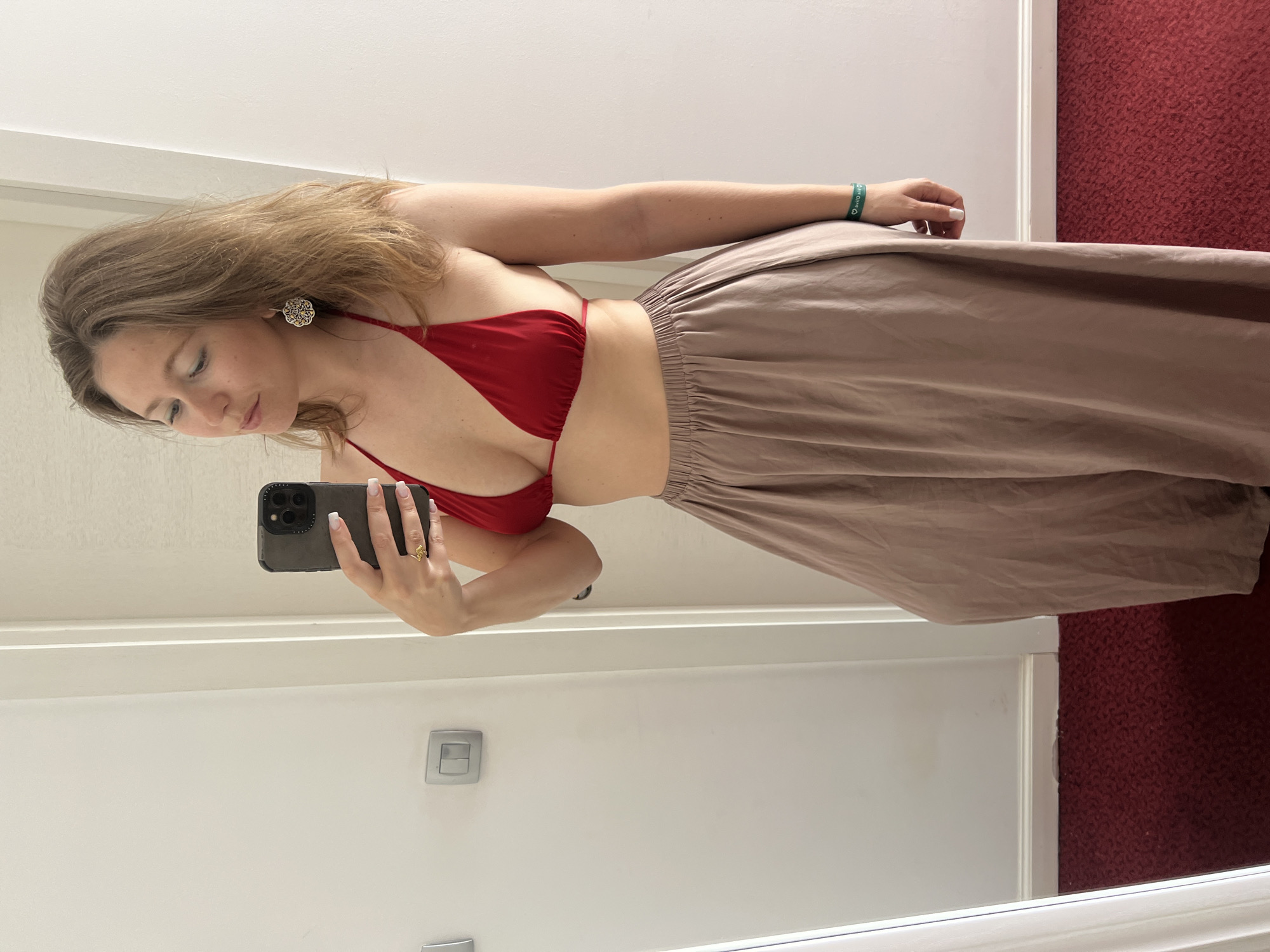 A woman is captured in a mirror selfie, showcasing her attire in an indoor setting. She has light brown hair and is looking downwards towards the smartphone she holds in her right hand. She is wearing a red bikini-style top and a flowing, light brown or beige wide-leg skirt or pants with a ruched elastic waistband.
On her left ear, she wears a distinctive, circular earring with an intricate design. A gold-colored ring is visible on a finger of her right hand, which is holding a dark-cased smartphone with a multi-lens camera array. Her fingernails are painted white with French tips. On her left wrist, she wears a green wristband that reads "ONE LOVE" alongside a small heart symbol.
The background features a plain white wall and a white wooden door frame. To the right, a section of red, patterned carpet is visible, suggesting the location is an interior space such as a room or corridor. A light switch with two gray buttons is mounted on the white wall. The overall lighting suggests an indoor environment.