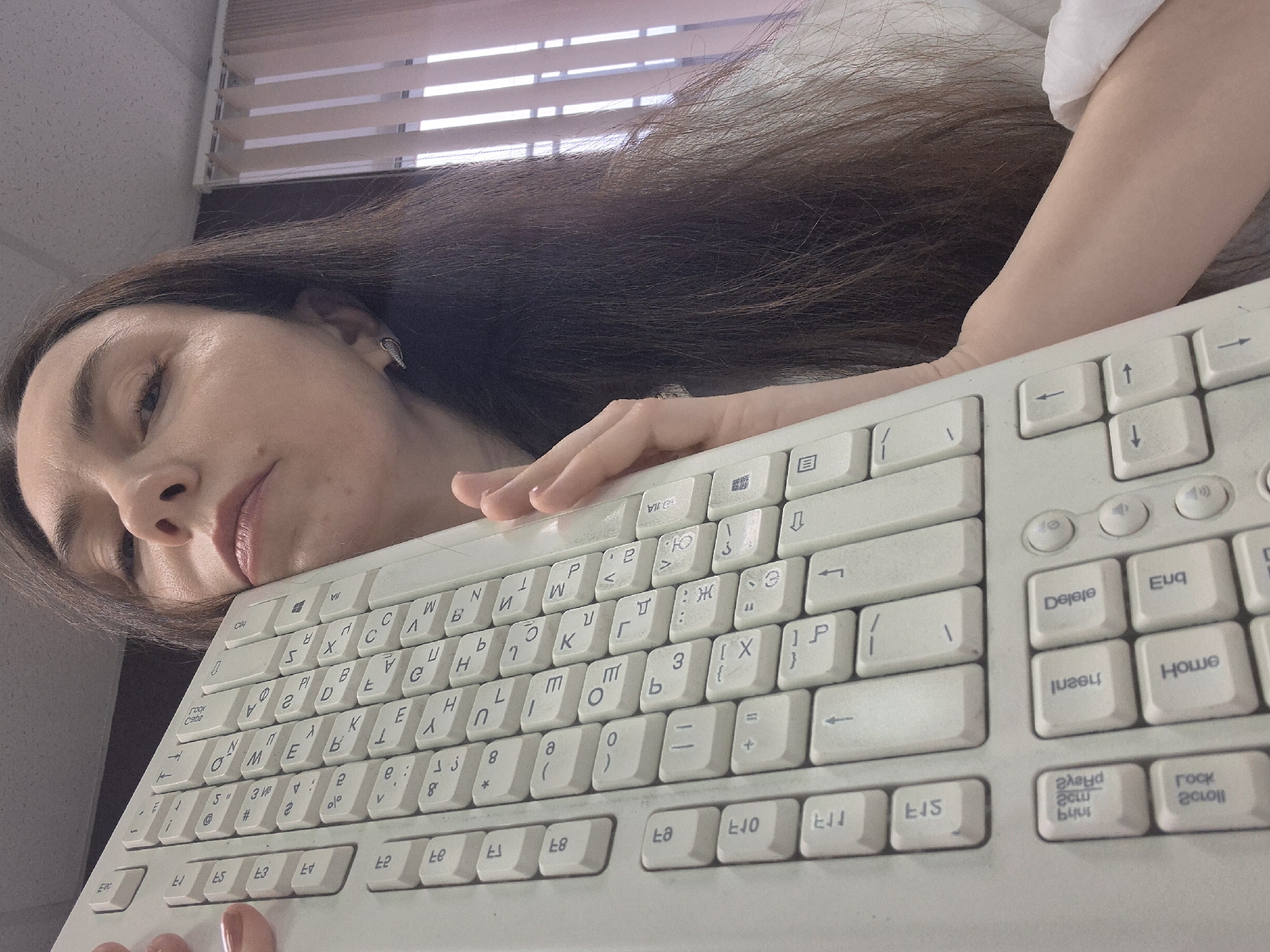 A woman with long, dark hair and fair skin is depicted resting her head on an old, off-white computer keyboard, viewed from a low-angle perspective. She is wearing a white top, and her eyes are partially closed, conveying a sense of tiredness or boredom. Her right arm and hand are also resting on the keyboard.
In the background, light filters through horizontal window blinds, indicating daylight. Above the blinds, a light-colored ceiling with acoustic tiles is visible, suggesting an indoor office or institutional setting.
The keyboard appears well-used and worn, featuring a layout with both Latin (QWERTY) and Cyrillic characters. Visible text on the keys includes standard English terms like "Delete," "Home," "Insert," "Scroll Lock," "Ctrl," "Shift," "Tab," and "Enter." Various function keys (e.g., "F5," "F8," "F10," "F11") are also discernible. Some text on the keys, such as "Delete" and "Home," appears slightly reversed due to the angle and wear.