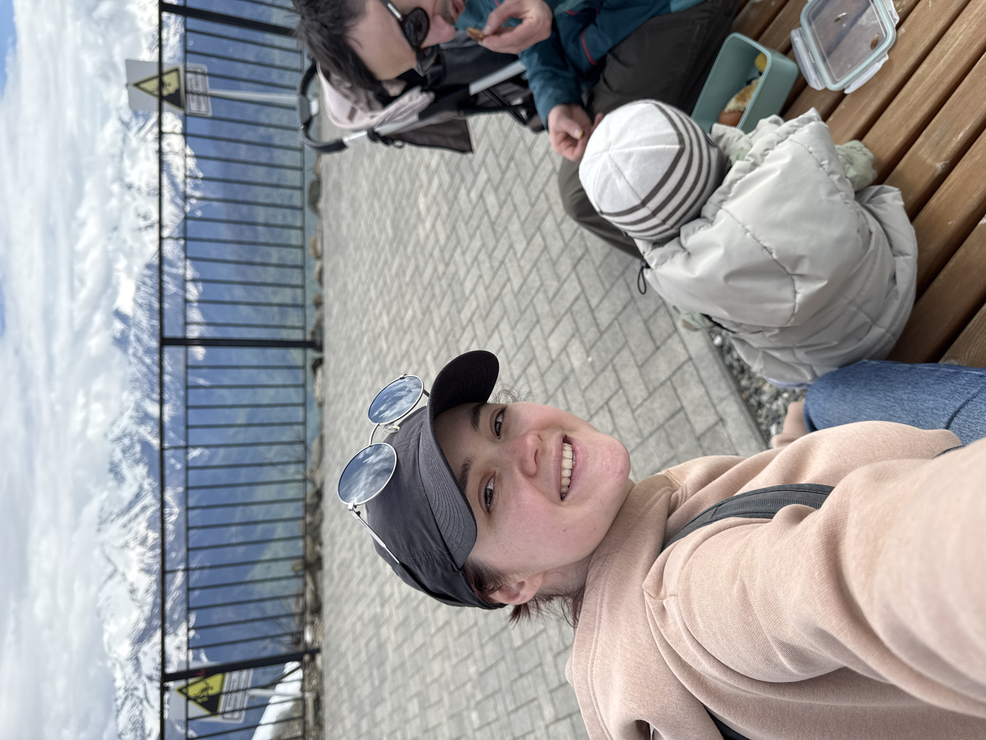 The image depicts three individuals at a high-altitude viewpoint in Estosadok, Russia, as suggested by the mountain range in the background. A person wearing a black cap and sunglasses is taking a selfie. Behind them, a person wearing sunglasses is eating. A child wearing a striped hat and a light-colored jacket sits on a wooden bench. The area appears to be paved with interlocking concrete blocks. The viewpoint has a metal railing, beyond which lies a mountainous landscape with snow-capped peaks. A warning sign is visible on the railing.