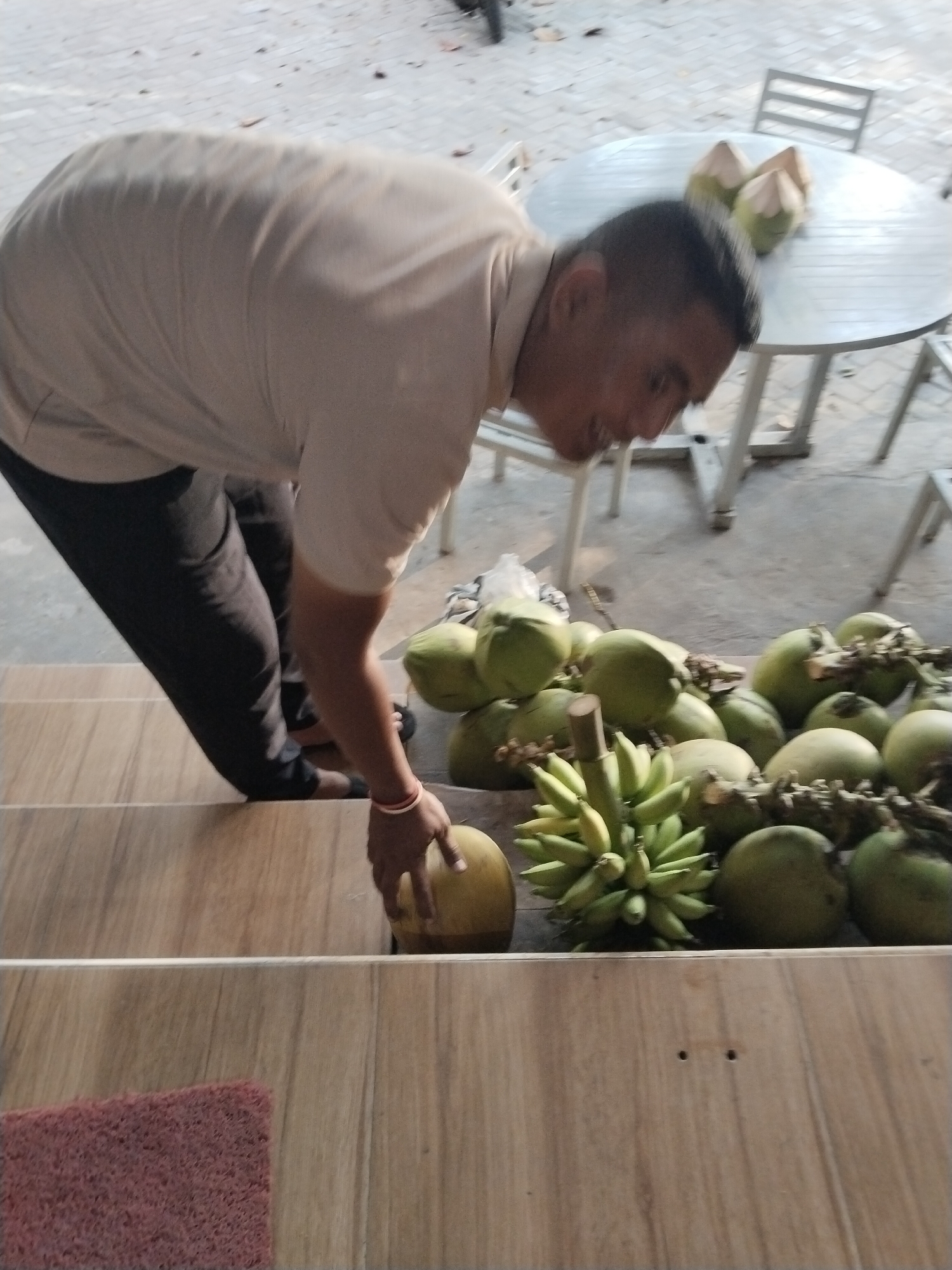 A man wearing a light brown shirt and dark pants is on stairs in Prigi, Indonesia. He is holding a large, round, brown fruit, possibly a coconut, in his right hand. In front of him, on the lower steps, is a pile of green coconuts and a bunch of green bananas. Behind him, on a table, are two more green coconuts. The ground is paved with light-colored bricks. The scene appears to be outdoors, possibly near a market or a roadside stall.