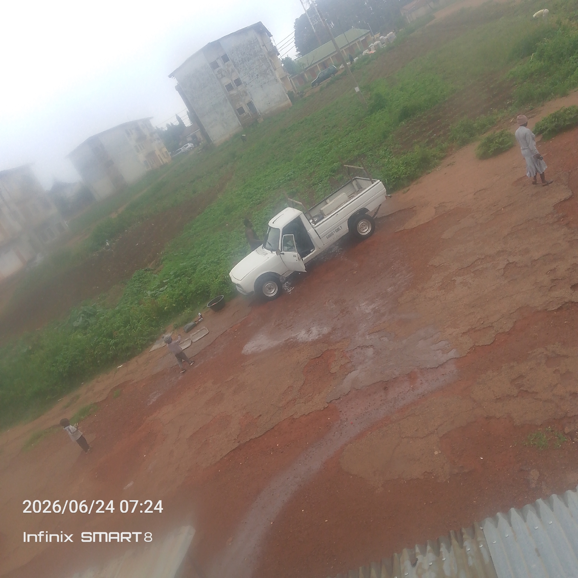 The image depicts a low-angle view of a dirt road in a somewhat hazy environment, possibly indicating fog or mist. In the foreground, a white pickup truck is parked with its driver's side door ajar. The truck has the text "GOLD ONLY" visible on its side. Near the truck, there are some white rectangular objects and a dark basin on the ground. A person in a light-colored robe, possibly a man, is walking away from the truck towards the right side of the frame. Further in the distance, several multi-story buildings are visible, some appearing weathered or unfinished, against a backdrop of green vegetation. A young child in a light-colored shirt and dark shorts is standing to the left of the truck, looking towards it. Another person is standing behind the truck. The overall scene suggests a casual, possibly rural or semi-urban setting. The date and time stamp "2026/06/24 07:24" and "Infinix SMART 8" are superimposed on the bottom left of the image.