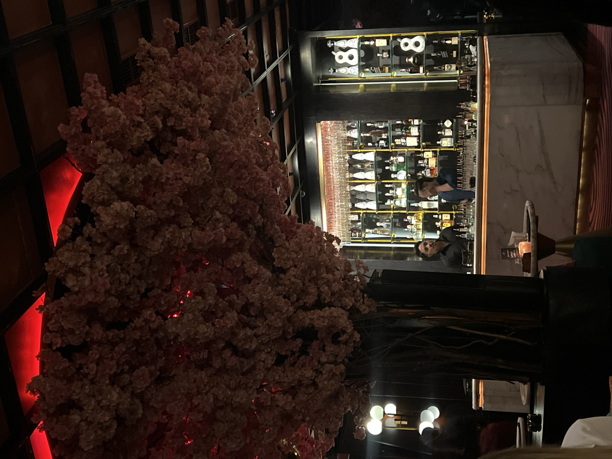 The image shows an interior view of a bar in North Kuta, Indonesia, taken from a table near an ornamental, pink-flowered tree-like structure with red illumination. The bar has a multi-tiered display of bottles and glasses, with two visible bartenders at work behind the counter. The bar counter is marble-like, and modern lighting fixtures are present. Part of the dining area, including tables and a partially visible person, is in the foreground. The scene is dimly lit.