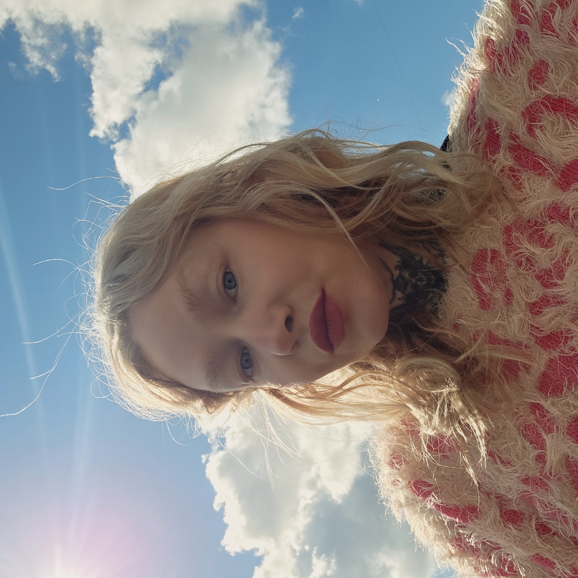 A low-angle portrait of a blonde woman with blue eyes and dark lipstick, taken outdoors. She is wearing a textured pink and white garment, possibly a sweater or jacket. Behind her is a bright blue sky with fluffy white clouds. The sun is visible in the lower left, creating a lens flare. The background suggests a location in Saint Petersburg, Russia, given the typical cloud patterns.