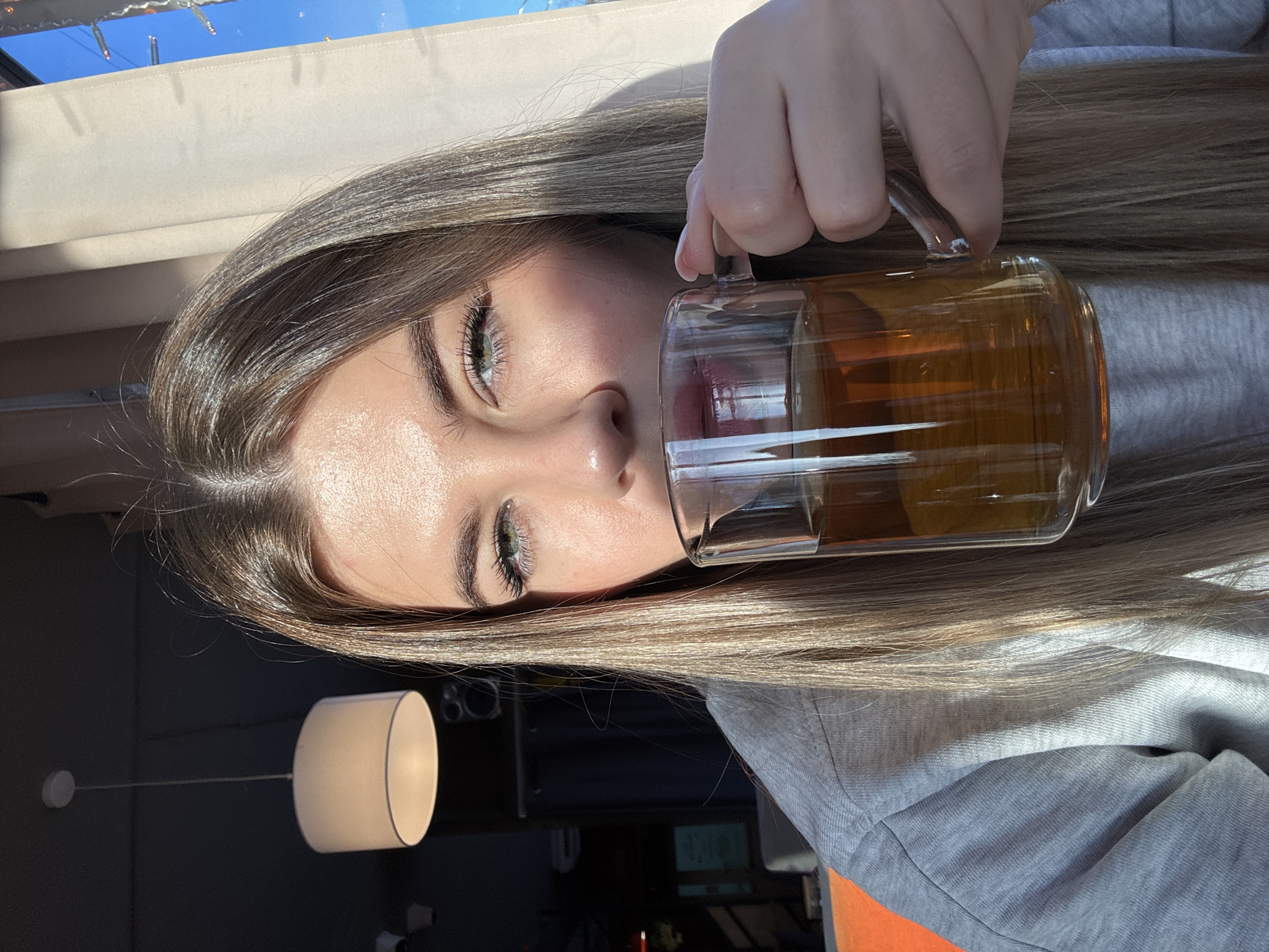 A female individual is depicted from the upper torso upwards, situated indoors within what appears to be a cafe or restaurant setting. She possesses long, straight, light brown hair and light-colored eyes, looking directly forward. The individual wears a light grey long-sleeved top. Her right hand holds a transparent glass mug, containing an amber-colored liquid, to her lips, indicating the act of drinking. Strong natural light, originating from the right side of the frame, illuminates the subject's right profile and portions of the interior. The background on the left is dark, displaying a suspended white drum-shade pendant light fixture and a dark wall-mounted speaker. On the right, a light-colored curtain or blind partially covers a window, through which a clear blue sky and a strand of decorative lights are visible. The precise location within Chaikovsky, Russia, cannot be determined from the visual information provided.