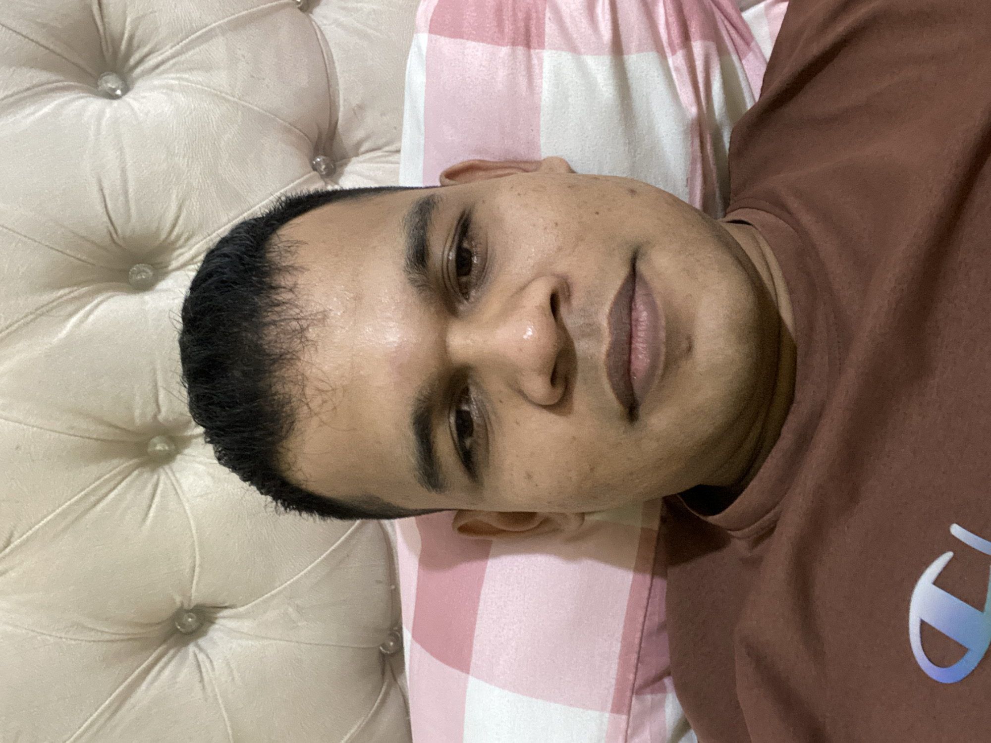 A man is lying down on a bed, with his head resting on a pillow. The background consists of a tufted headboard. The man is wearing a brown t-shirt with a logo on the left side of his chest. The pillow beneath his head has a pink and white checkered pattern. The image appears to have been taken indoors. The man is facing directly towards the camera.