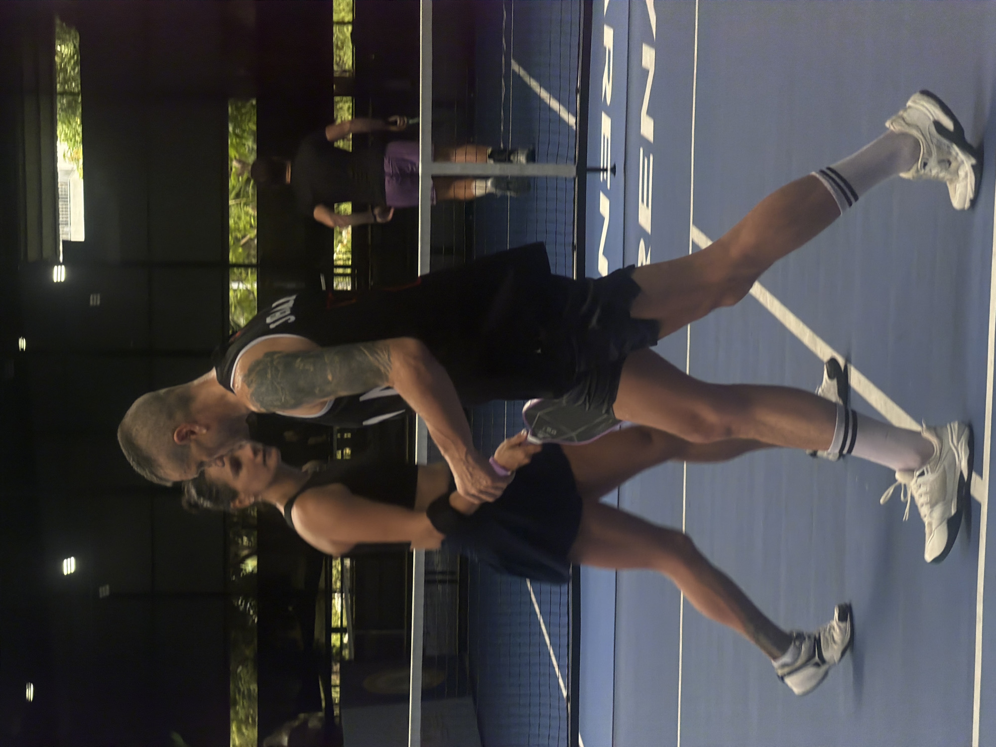 The image depicts a tender moment between a man and a woman on a blue padel court, likely in Nam Nha Trang Ward, Vietnam. The scene appears to be set in the evening or at night, indicated by the dark background, artificial lighting, and illuminated green foliage visible outside the court.
In the foreground, a man with a shaved head and tattoos on his left arm, wearing a dark tank top and shorts, stands facing a woman. His hands are placed around her waist/hips. The woman, dressed in a dark athletic top and skirt, has her hands on his arms as she looks up at him. Their proximity suggests an intimate or affectionate moment. Both are wearing athletic shoes with white laces and socks, the man's socks featuring black stripes.
In the blurred background, two other individuals are visible, one holding what appears to be a padel racket, indicating that a game is either in progress or has recently concluded. The court itself is blue with clear white boundary lines and is divided by a net. Prominently visible on the blue court surface, beyond the net, are large white letters spelling "ARENA" (or possibly "REN" as part of a larger word). The overall atmosphere combines a personal, emotional interaction with a dynamic sports setting.