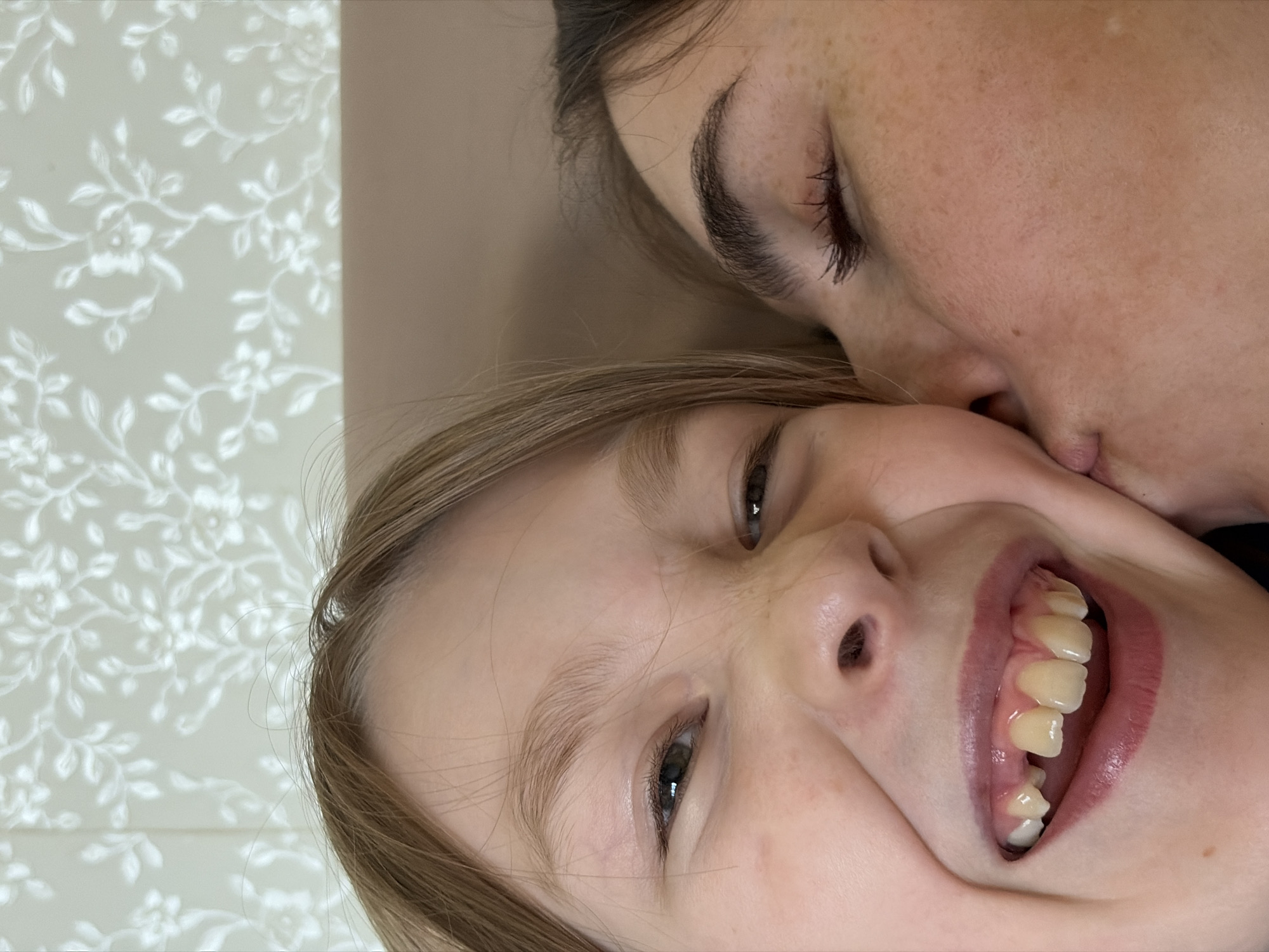 A close-up, off-center image shows a child's face and a portion of an adult's face. The child, with fair skin and blonde hair, is grinning with visible teeth. The adult, with dark hair, is positioned such that their cheek is near the child's face. The background consists of a light-colored wall with a floral pattern and a plain, light-brown surface. The location of the image is not deducible from the photograph.