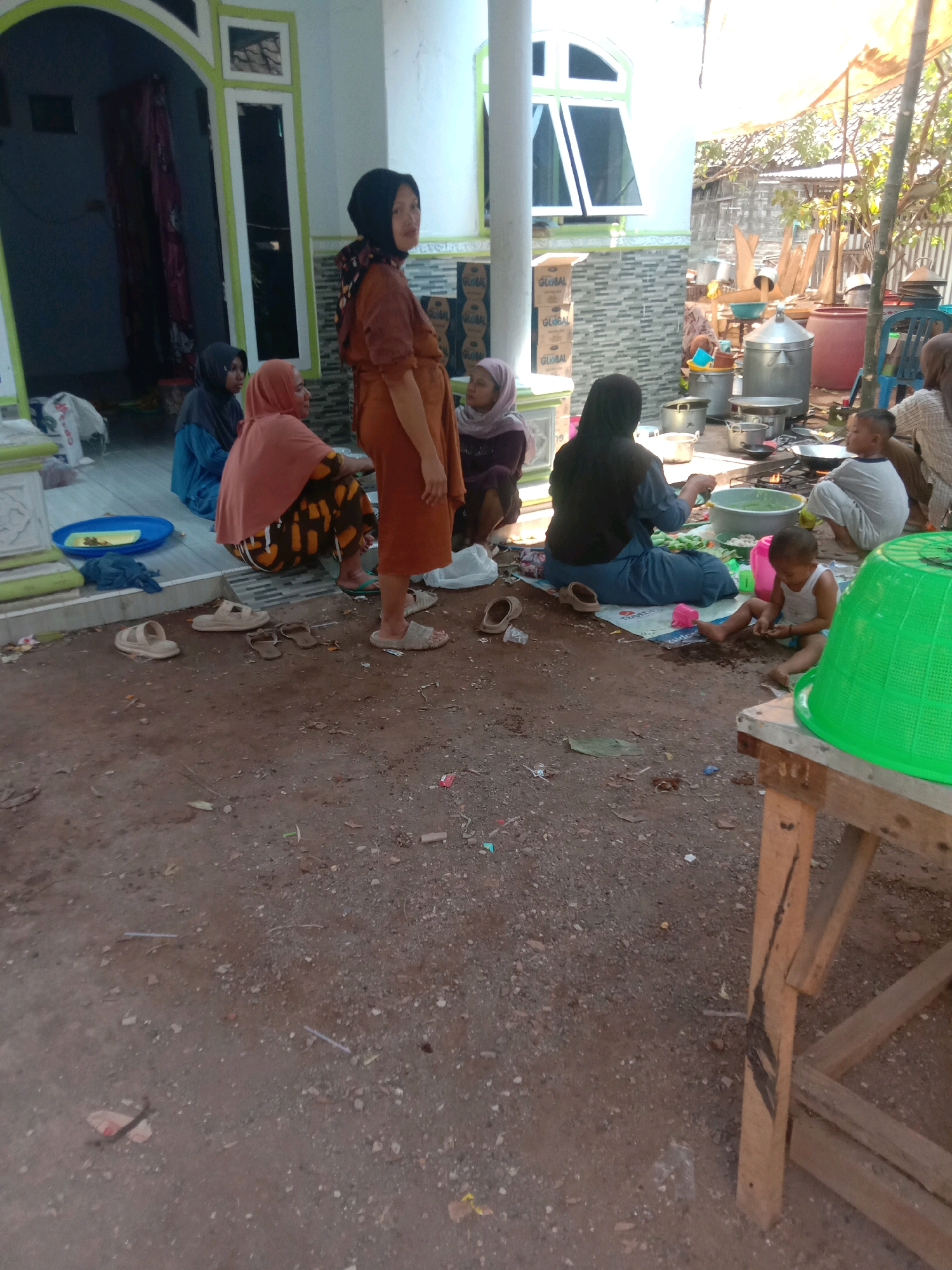 In Karang Anyar, Indonesia, a group of people, predominantly women and children, are gathered outdoors near a building with a white facade and green trim. The scene appears to be a communal gathering or event, possibly related to food preparation. Several individuals are seated on the ground, some wearing hijabs. One woman stands, wearing an orange dress and headscarf, looking towards the camera. Another woman in a blue headscarf and blue top is visible. A young child in a white tank top is sitting on the ground near a large green basket. In the background, various cooking pots and utensils are laid out, suggesting food is being prepared. The ground is dirt and gravel, with scattered debris. The environment is sunny, with trees and a corrugated roof visible in the distance. The overall atmosphere suggests a casual, everyday setting.