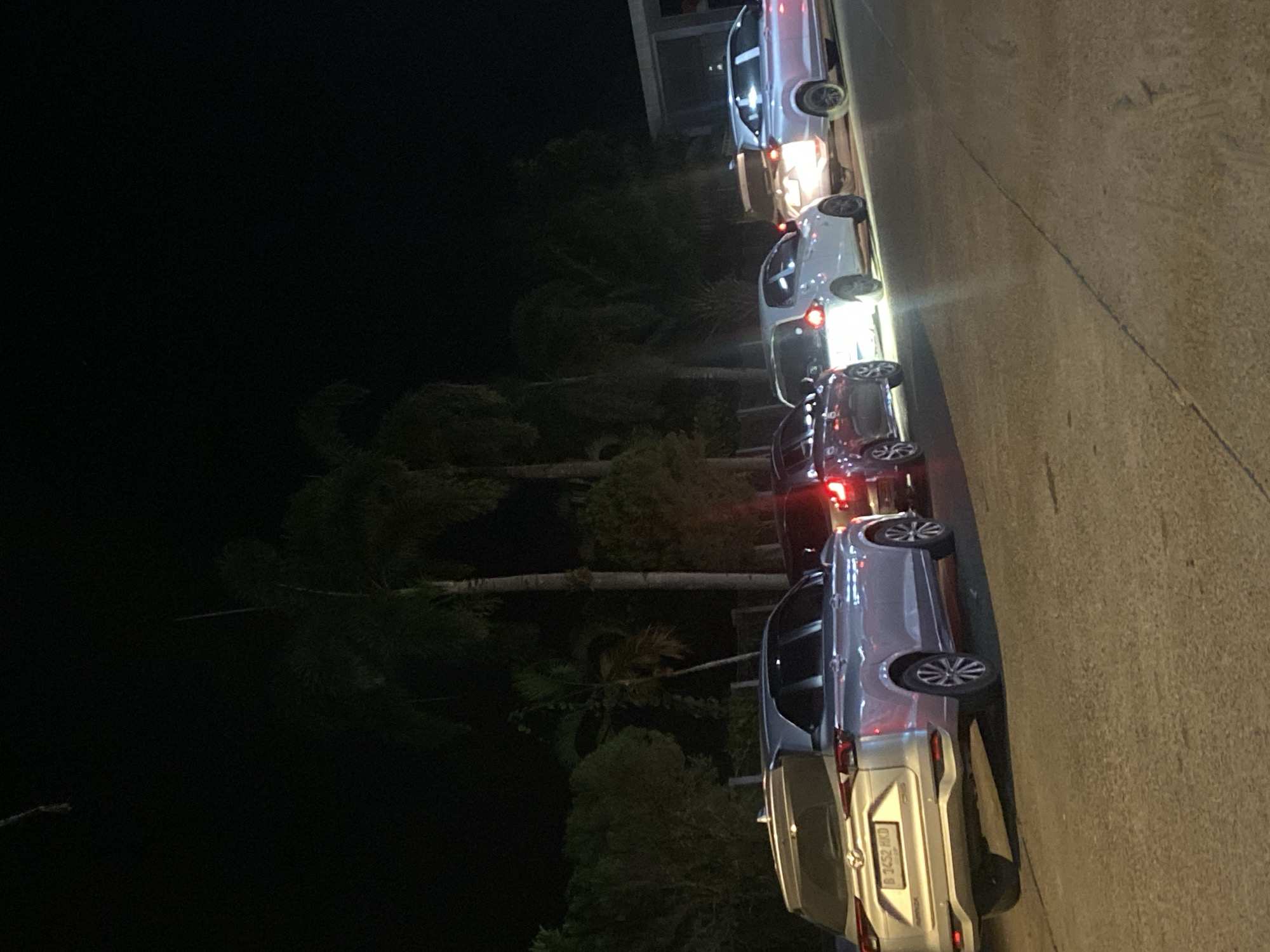 A line of vehicles is parked on a paved surface at night, with palm trees and vegetation in the background. The scene appears to be in Gambut Barat, Indonesia. The most prominent vehicle in the foreground is a silver SUV with its rear facing the camera, displaying a license plate that reads "B 3452 HKD". Several other cars are visible behind it, some with their headlights or taillights illuminated. The lighting conditions suggest it is nighttime, with artificial lights casting reflections on the vehicles and road. The environment is dark, with the sky being black and the trees silhouetted against it.