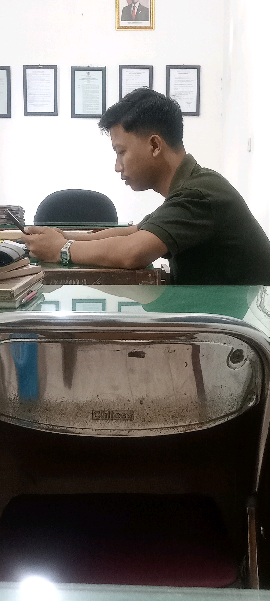 A young man with short black hair, wearing a dark green collared shirt, is seated at a desk and looking down at a smartphone in his hands. His left arm is resting on a stack of books, and he has a silver watch on his left wrist. Behind him, on a white wall, are several framed documents and a portrait of a man in a gold frame. The desk surface is made of glass, and underneath it, a chrome-colored chair with a dark seat is visible, bearing the embossed name "Chitose." The scene appears to be an indoor office environment.