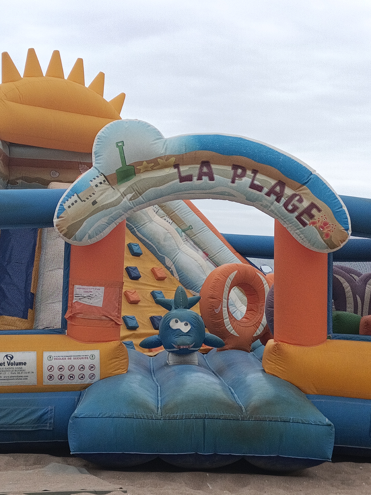 An inflatable beach-themed attraction, likely for children, stands on a sandy surface under an overcast sky. The structure features a large, arched entrance adorned with the text "LA PLAGE" and illustrations of a shovel, sand, and a crab. Beneath the arch, a slide is visible, and to the left, a climbing wall with colorful handholds. A blue, cartoonish sea creature figure is positioned in the center of the inflatable. On the left side of the structure, a label with text including "Street Volume" and "REGLES DE SECURITE" (Safety Rules) is attached. The inflatable is predominantly blue and orange, with yellow elements. The overall scene suggests an outdoor recreational area.
