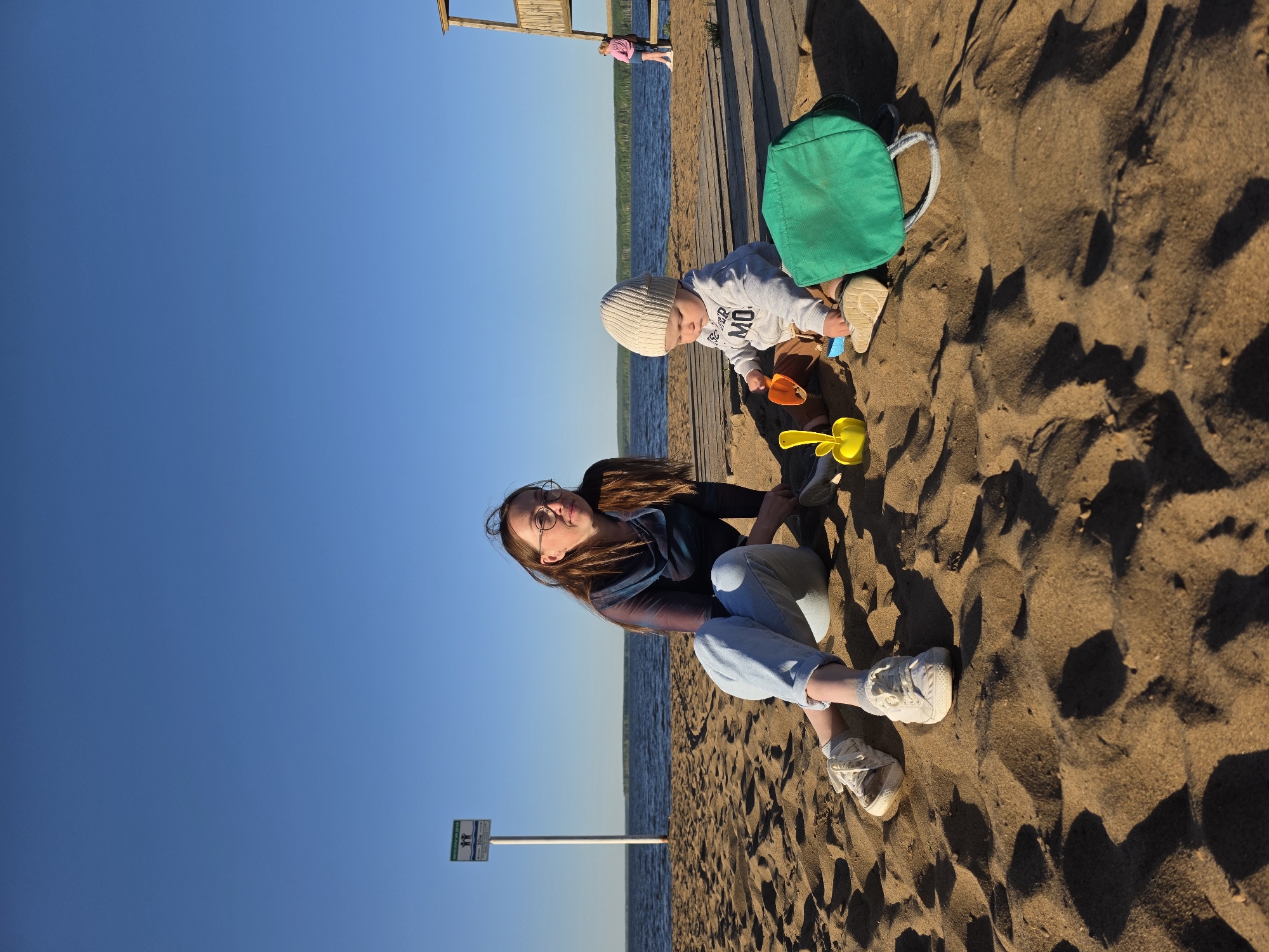 A woman and a baby are playing on a sandy beach. The woman, wearing glasses, is sitting on the sand in a navy jacket and light blue jeans. She is smiling and looking towards the camera. The baby, wearing a white beanie and a gray sweatshirt with text, is playing with sand toys near a green backpack. The beach leads to a body of water. The background includes a wooden boardwalk and a sign on a pole. The location is Chaikovsky, Russia.