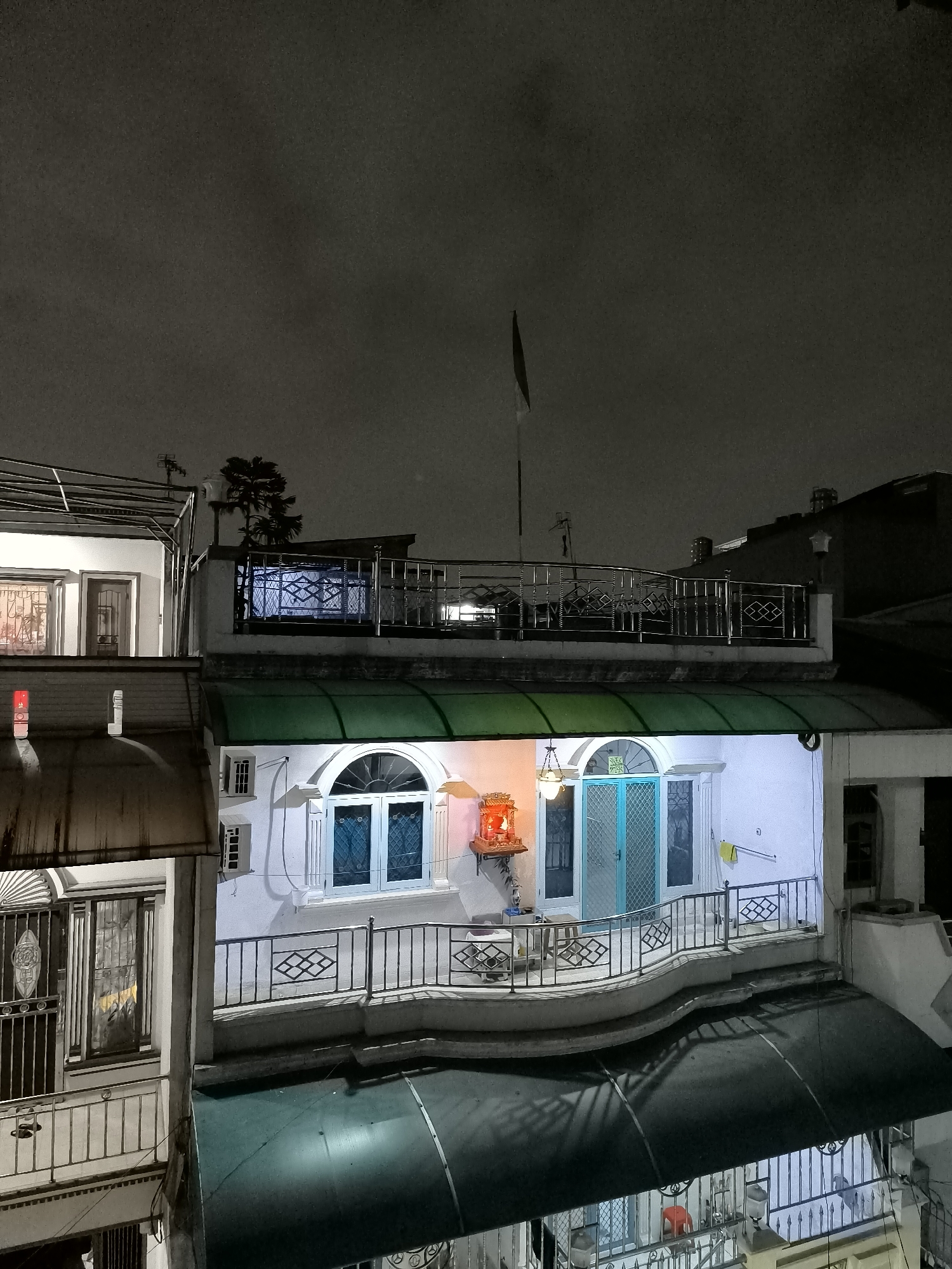 The image shows a multi-story building at night, possibly in North Jakarta, Indonesia, with various architectural features and lighting. On the uppermost level, a metal railing surrounds a flat roof area. Below this, the second floor features large arched windows with blue panes and a balcony with a metal railing. A green awning extends from the second floor. The third floor has windows with decorative metal bars and what appears to be a corrugated metal roof. The lighting suggests an urban environment, with streetlights and interior lights illuminating parts of the building. The overall scene is dark, with the sky appearing overcast or nighttime. There are no people visible in the image.