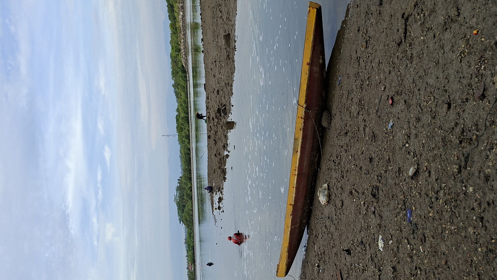 The image depicts a muddy shoreline at low tide, with a long, narrow canoe resting on the bank in the foreground. The canoe is painted red and yellow. In the shallow water, several individuals are engaged in activities, with one person in a red shirt partially submerged, appearing to be fishing or collecting something. Further in the distance, other figures are visible on the exposed mudflats. The background features lush green vegetation lining the opposite bank of a body of water, with mountains and a transmission tower visible on the horizon under a cloudy sky. The scene suggests a coastal or riverine environment in the city of Lhokseumawe, Indonesia, possibly during a low tide fishing or gathering activity. The shoreline is littered with debris, including plastic waste and shells, indicating a natural environment that is also impacted by human activity and waste.