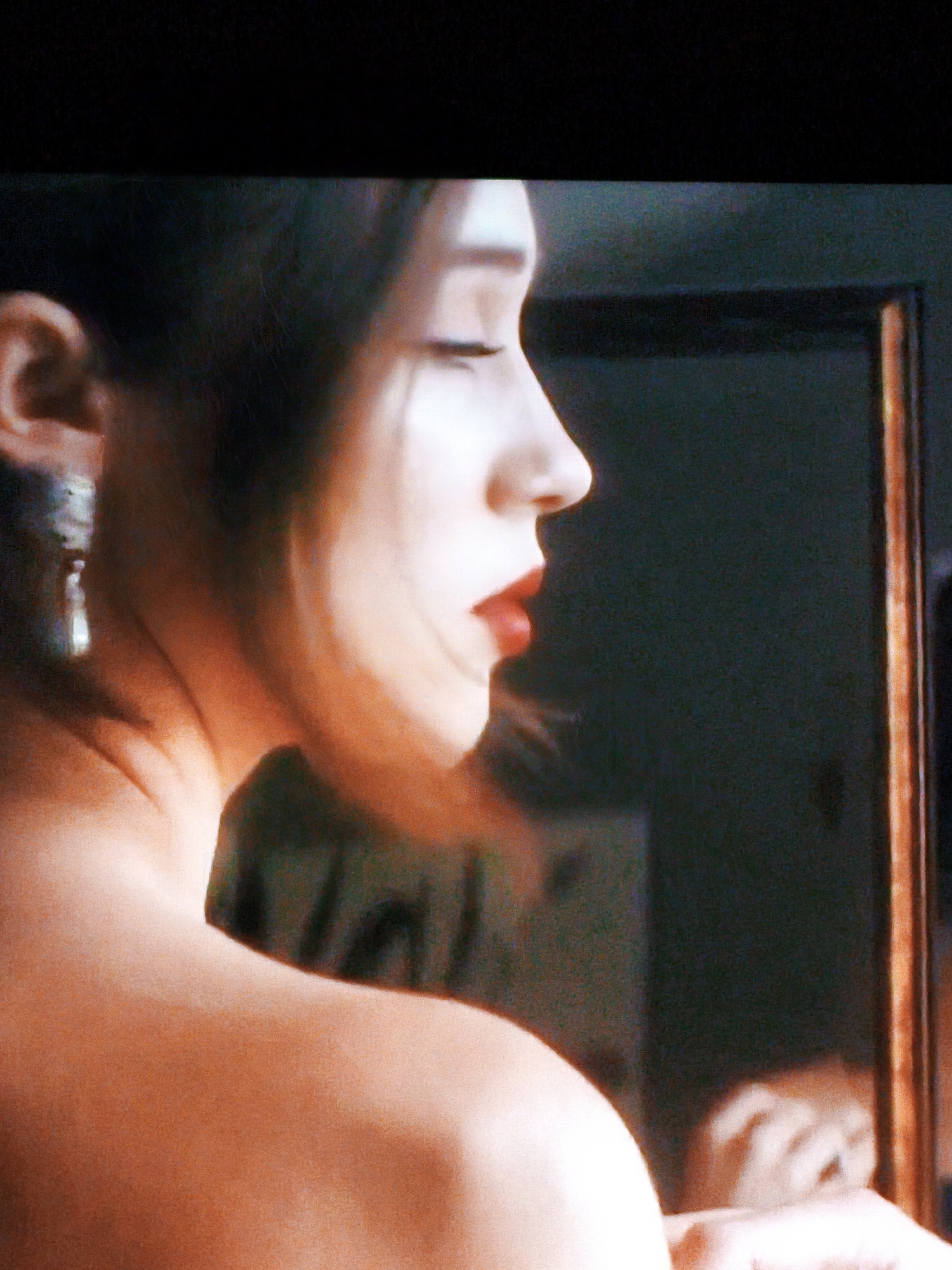 The image is a close-up, profile view of a woman's face, with her head tilted slightly upwards. She has dark hair, parted on the left, and is wearing bright red lipstick. A prominent earring, a silver dangle design, is visible on her left ear. Her bare shoulder and upper back are also in view, illuminated by warm lighting. In the background, a dark surface, possibly a wall, is visible, with a blurred reflection of a mirror frame on the right side. The overall lighting is dim and atmospheric, with a reddish-orange hue dominating the skin tones. The location is not discernable from the image.