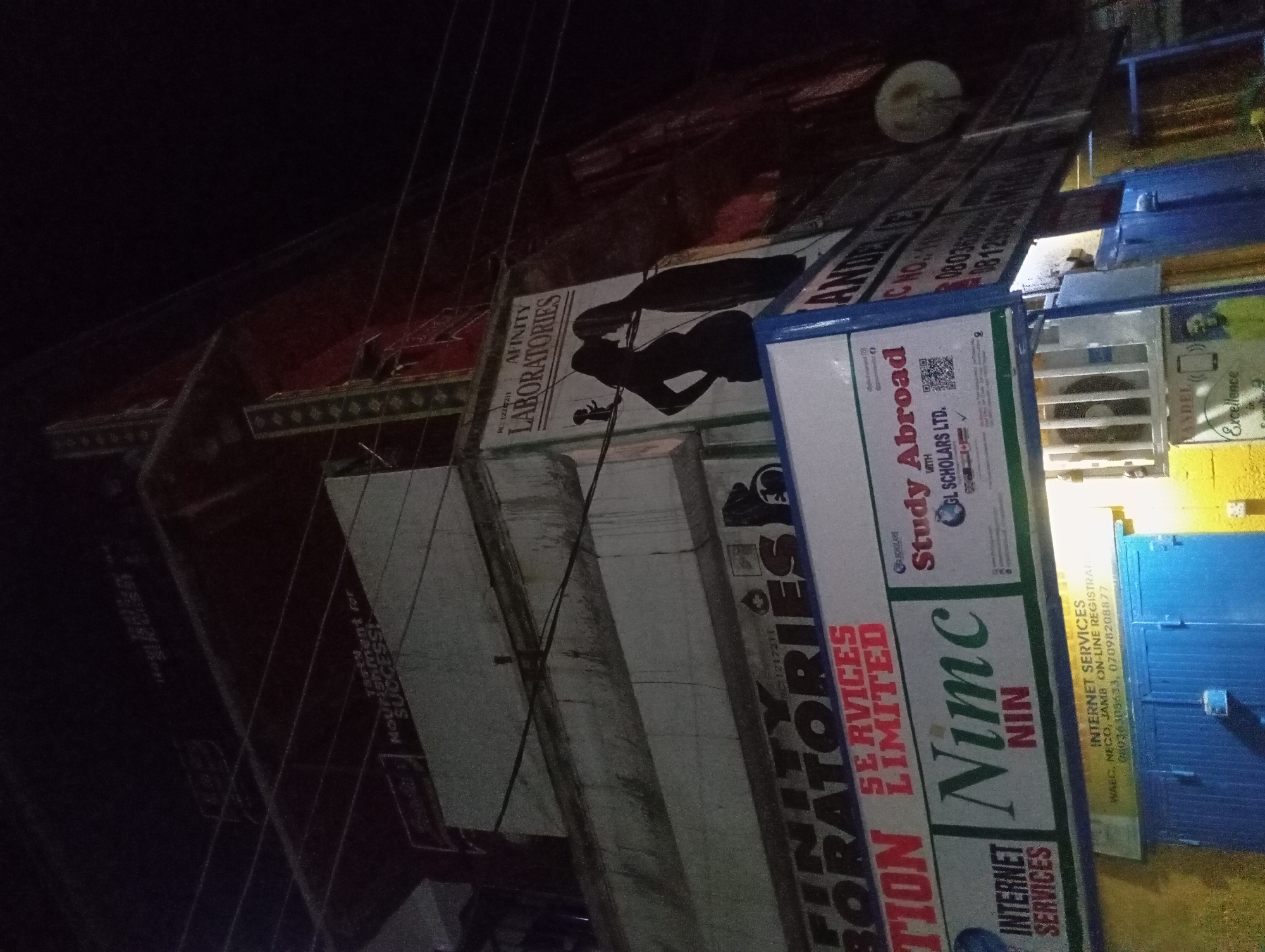 An exterior nighttime shot of a multi-story building in an urban setting. The building has multiple signs and advertisements. The main focus is a sign for "AFFINITY LABORATORIES" with a silhouette of a pregnant woman and a man kissing. Below this sign, another sign reads "NIMC NIN" and "INTERNET SERVICES." Further down, there is a sign for "Study Abroad" with "GL SCHOLARS LTD." written on it. Additional signs and wires are visible across the building. The lower part of the image shows a closed blue shuttered doorway with a yellow frame. The lighting is dim, suggesting it's night.